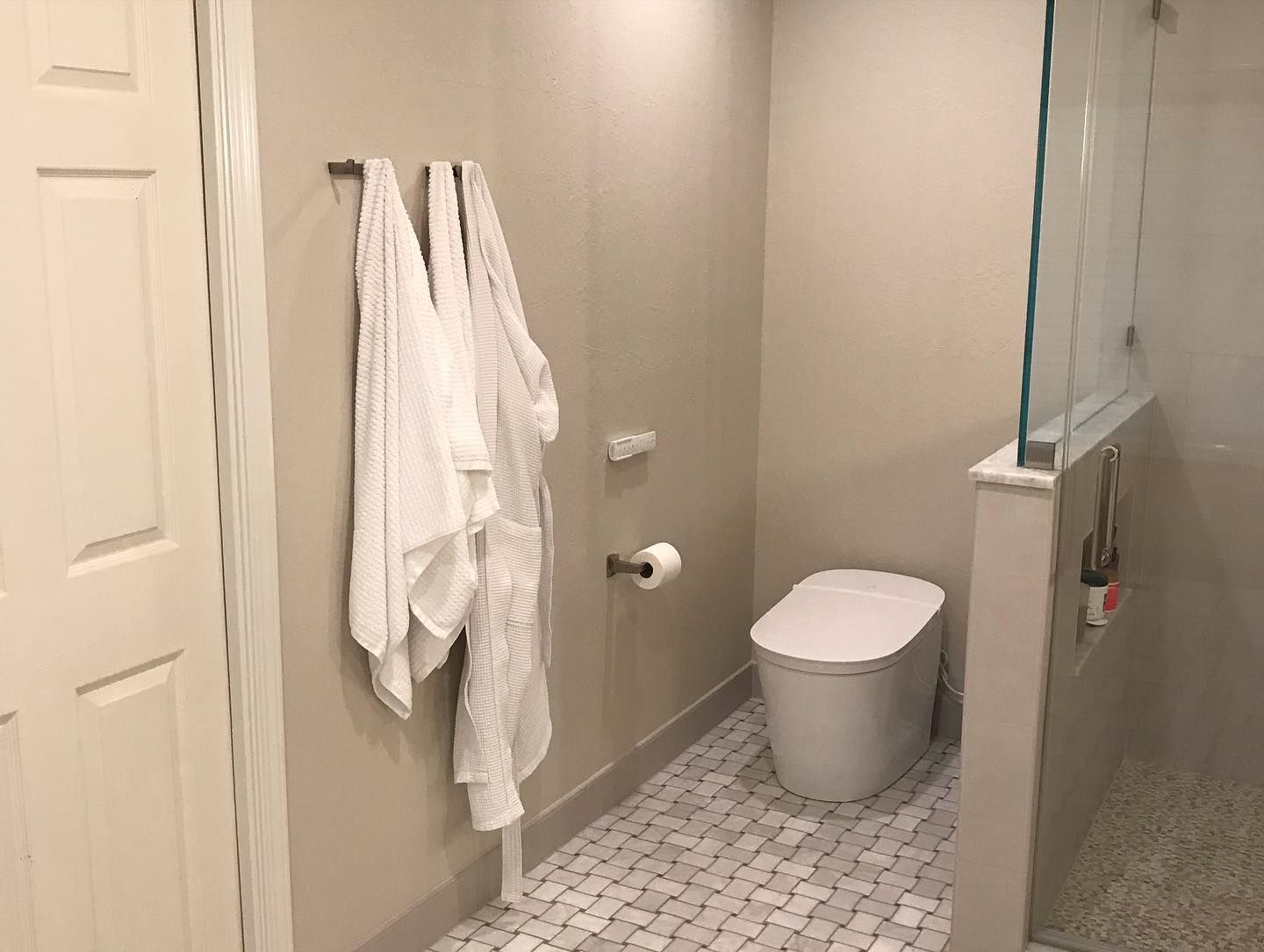 A small, neutral-toned bathroom featuring a toilet, a shower with a glass partition, and white towels hanging on the wall.