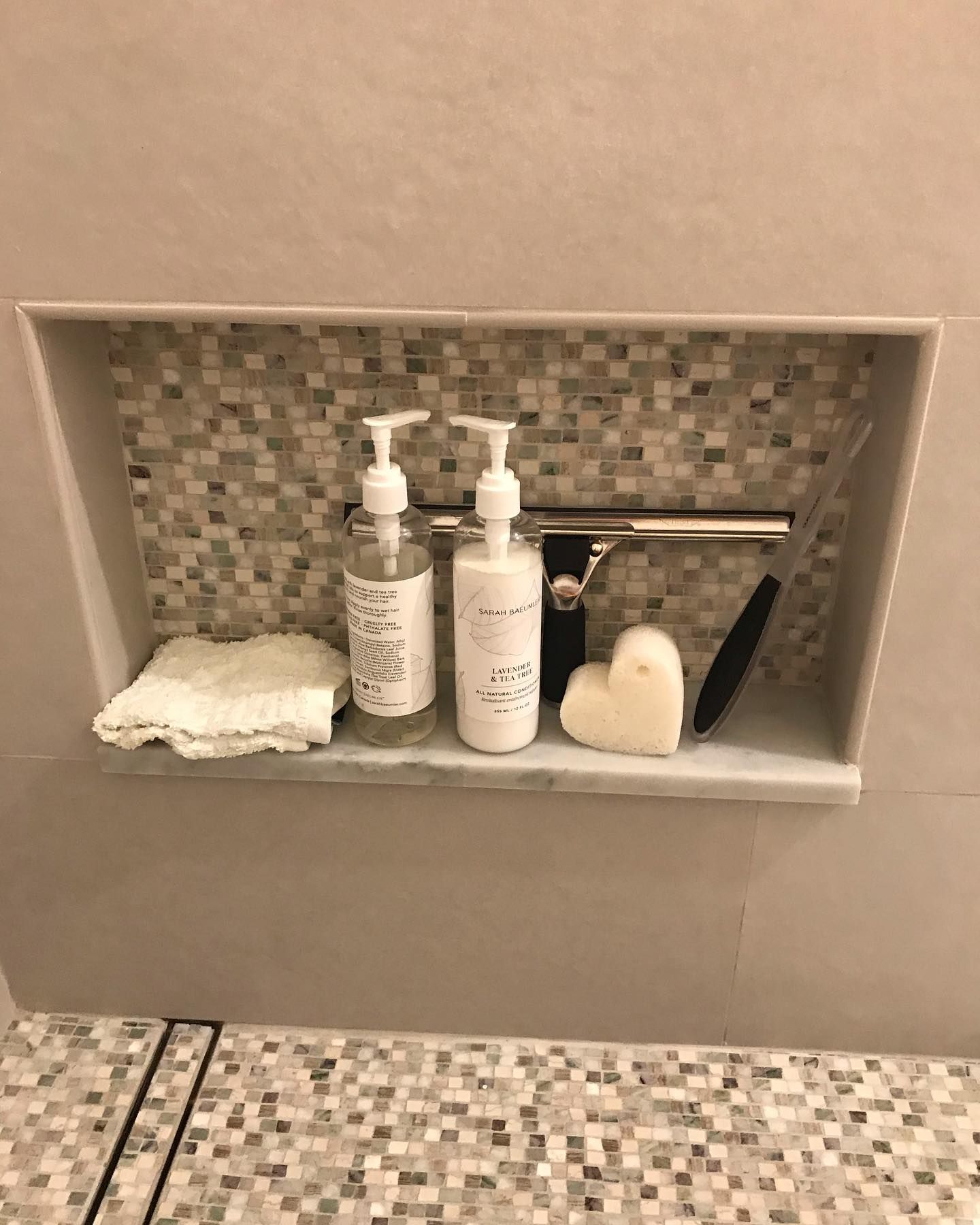 A tiled shower wall niche holding two pump bottles, a squeegee, a folded white washcloth, and a heart-shaped bar of soap.