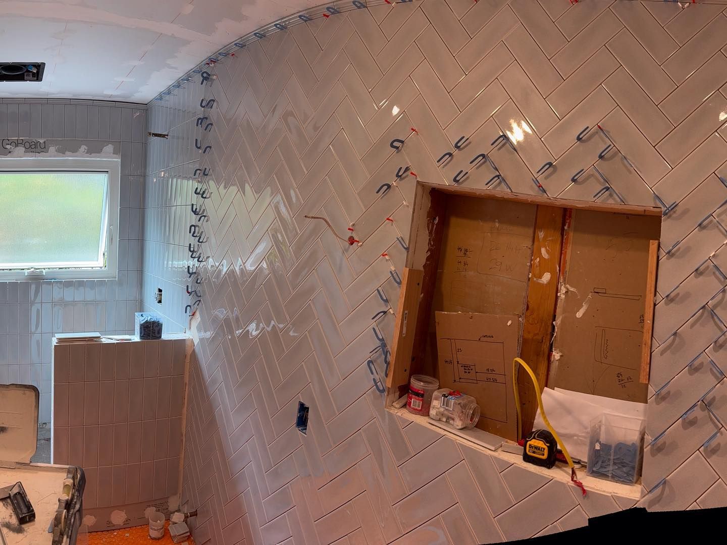 A half-tiled wall in a room under construction with a white herringbone pattern and a rectangular recessed nook.