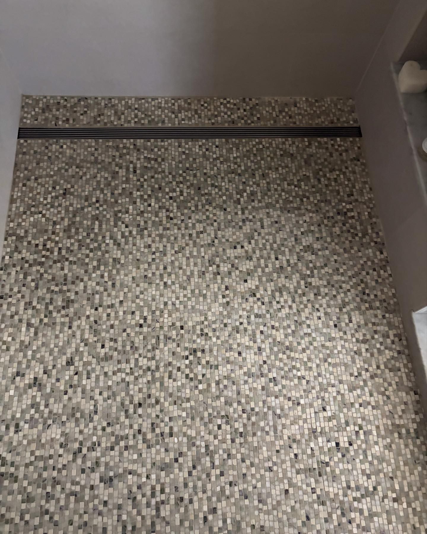 A shower floor covered in small, multi-toned gray and white square tiles with a linear black drain across the back.