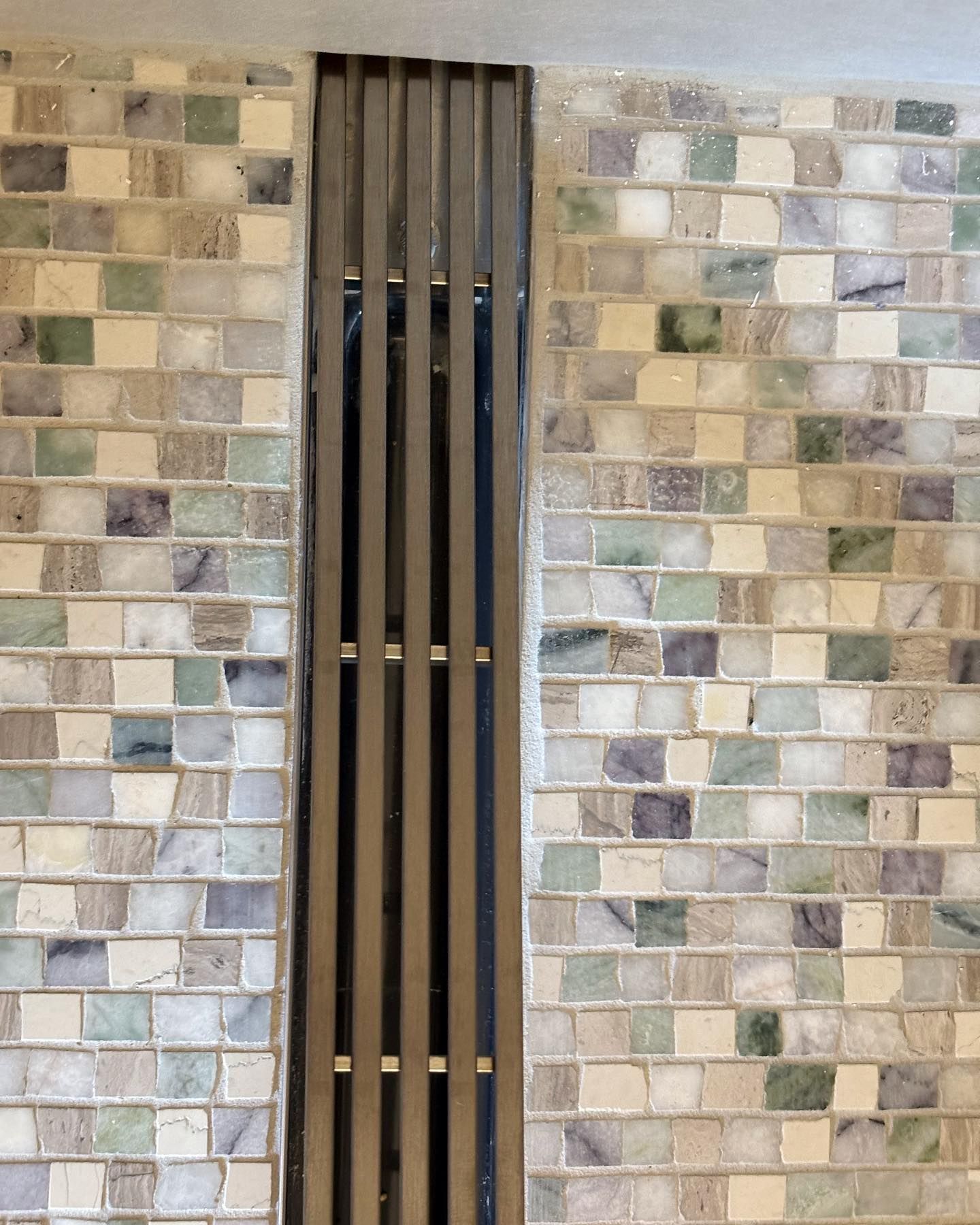 A vertical wooden slat screen is set between two walls covered in small, multi-colored, light-toned mosaic tiles.