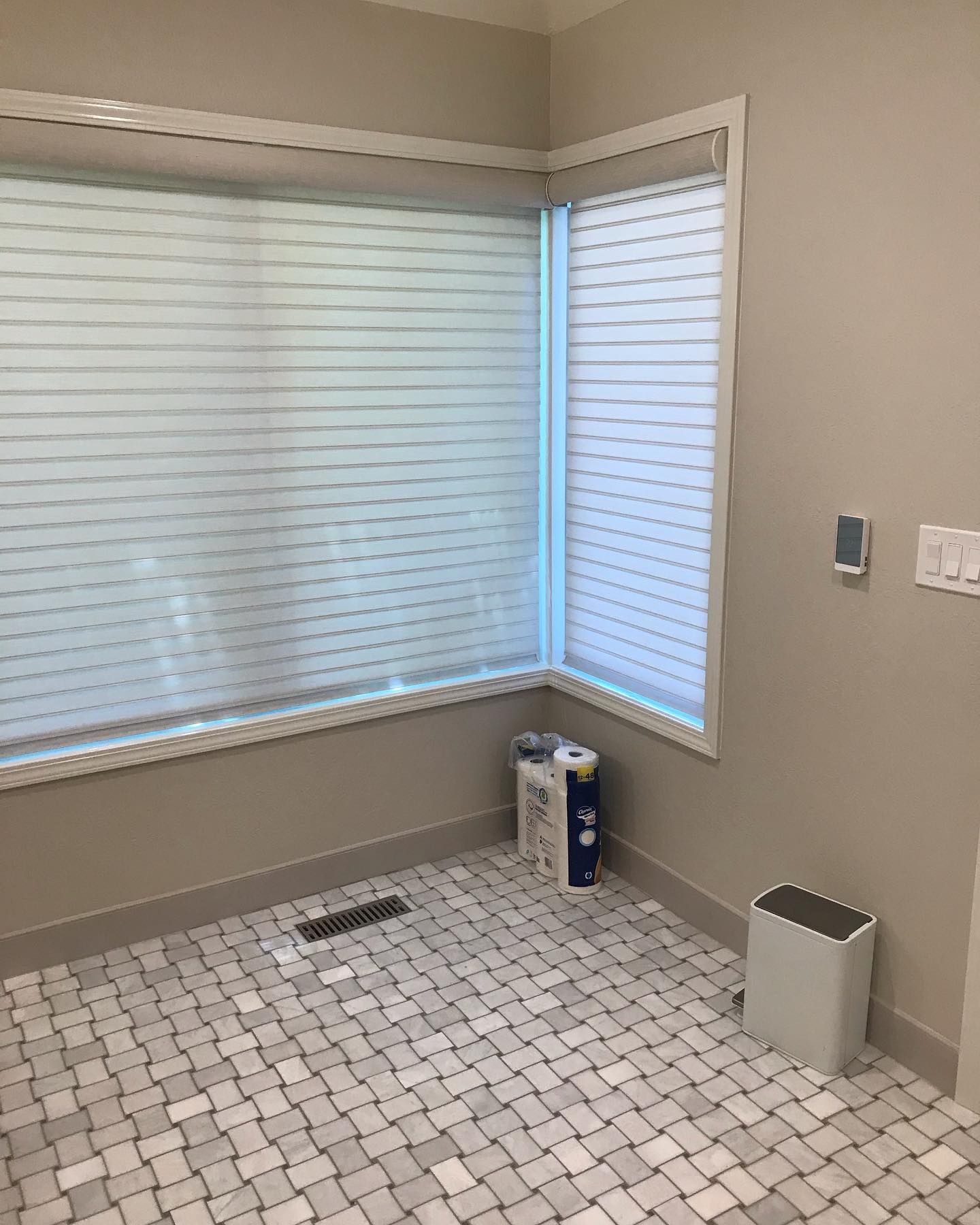 Corner view of a room with light-colored walls, large windows with horizontal blinds, tiled flooring, and a small air unit.