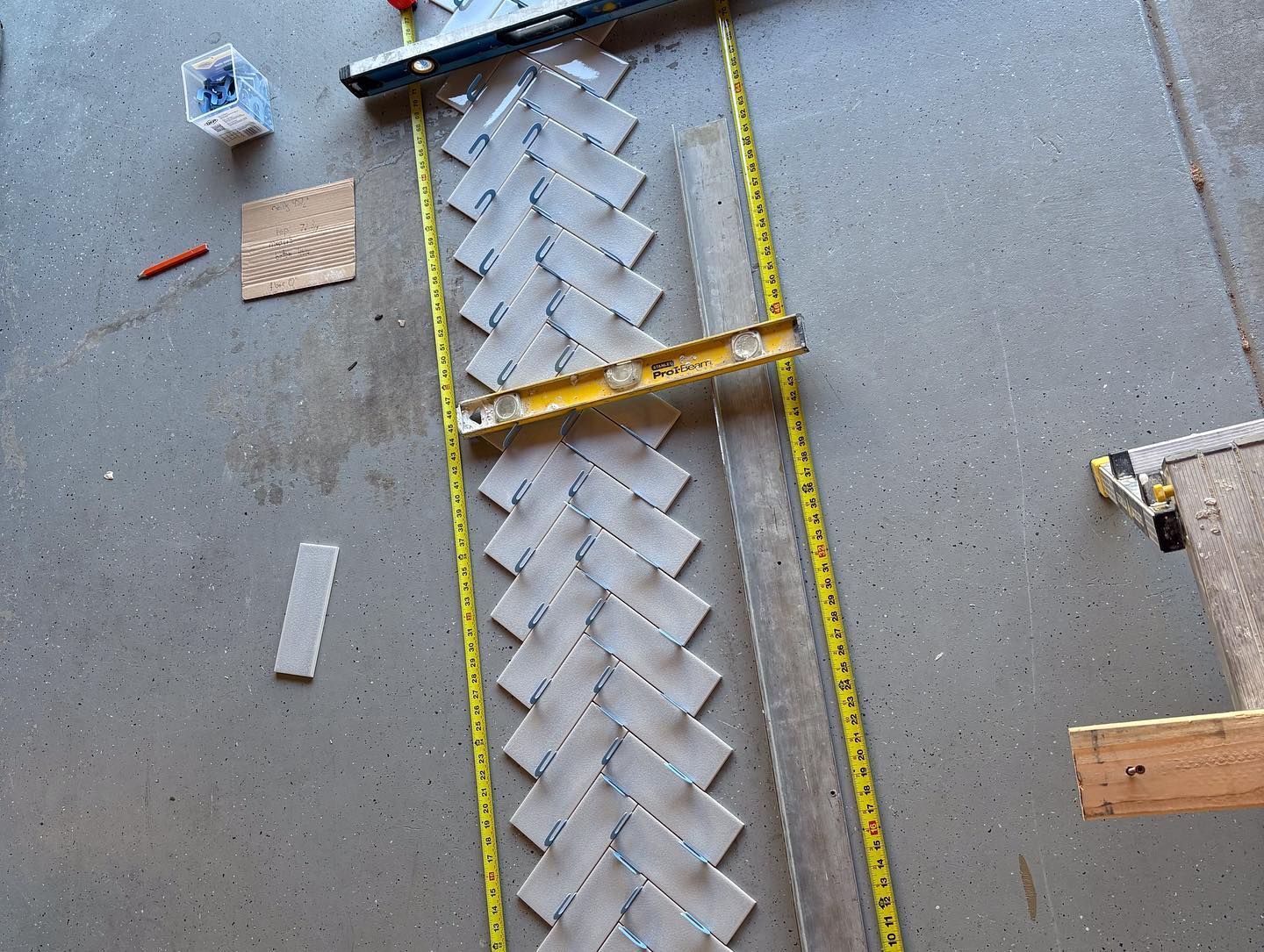 Light grey rectangular tiles are laid in a herringbone pattern on a concrete floor, framed by measuring tapes and a level.
