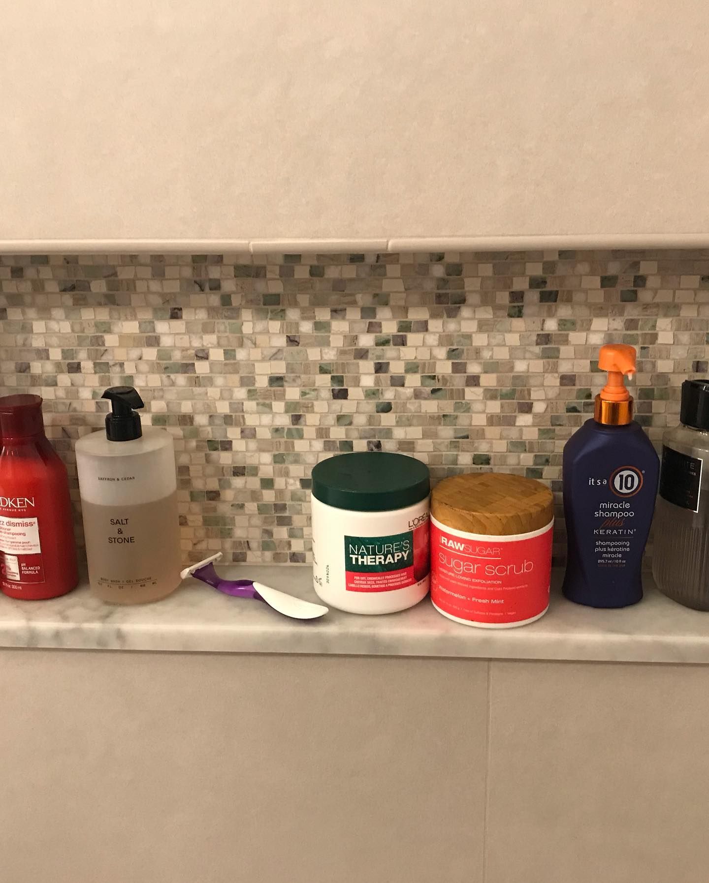 A row of various bottles and jars of hair and bath products sit on a marble shower ledge against a mosaic tile wall.