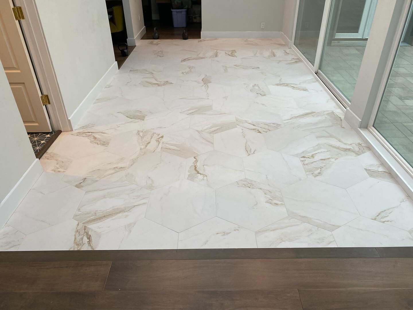 A transition from dark wood flooring to light, marbled, hexagonal tile flooring in a bright residential interior.