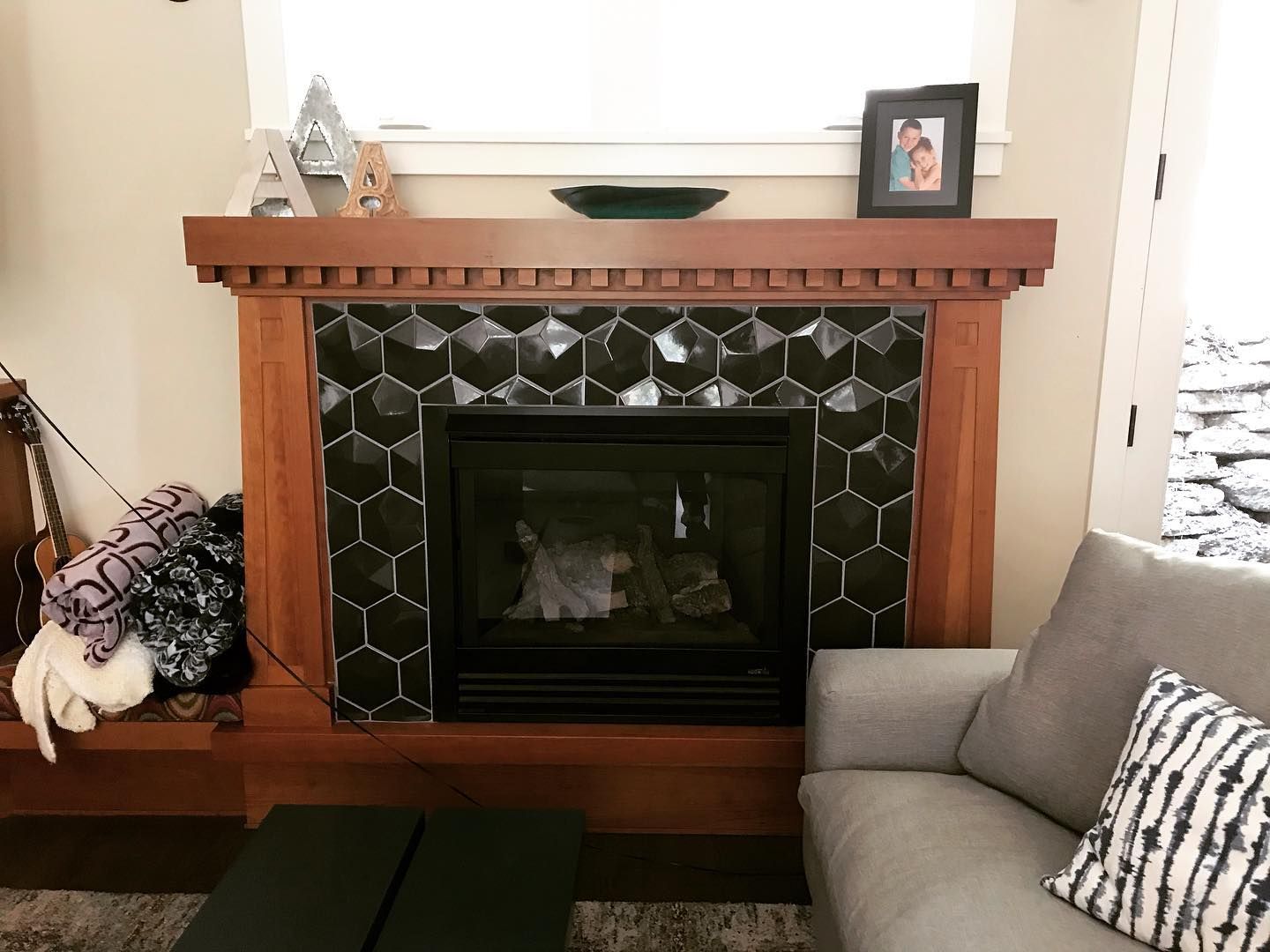 A fireplace with a warm wood mantle and black hexagonal tile surround, featuring decorative items and a nearby sofa.