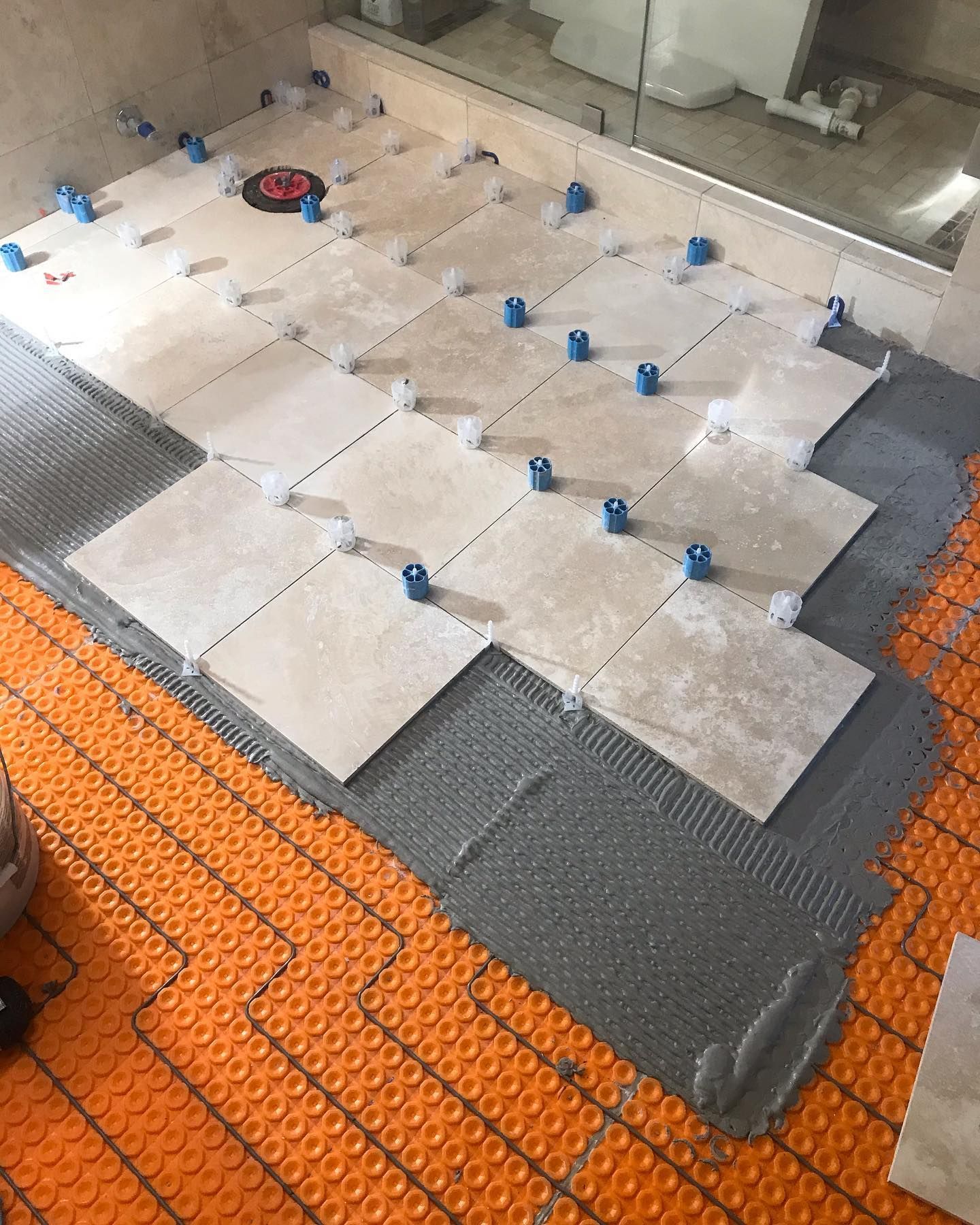 Light-colored square tiles are being laid over an orange floor heating membrane in a bathroom during renovation.