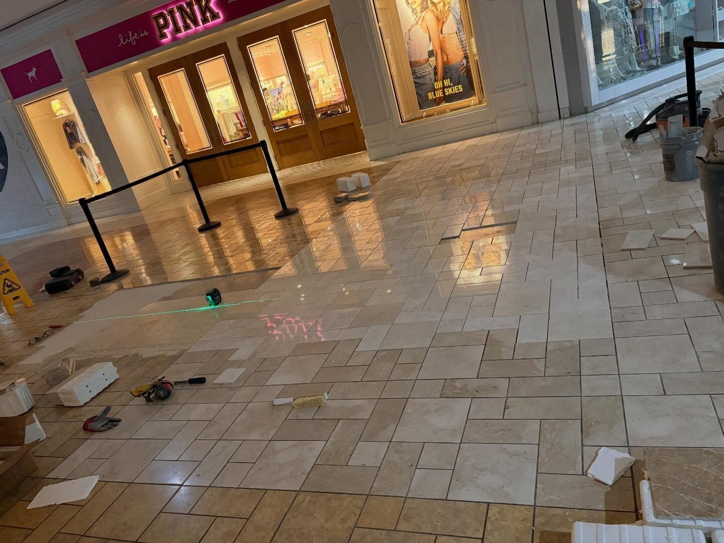 Construction work in a shopping mall near a storefront, with partially replaced light-colored stone floor tiles.