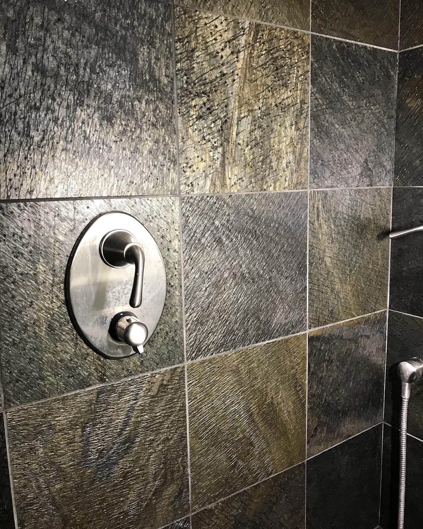 A stainless steel shower faucet handle mounted on dark, textured, multicolored stone wall tiles.
