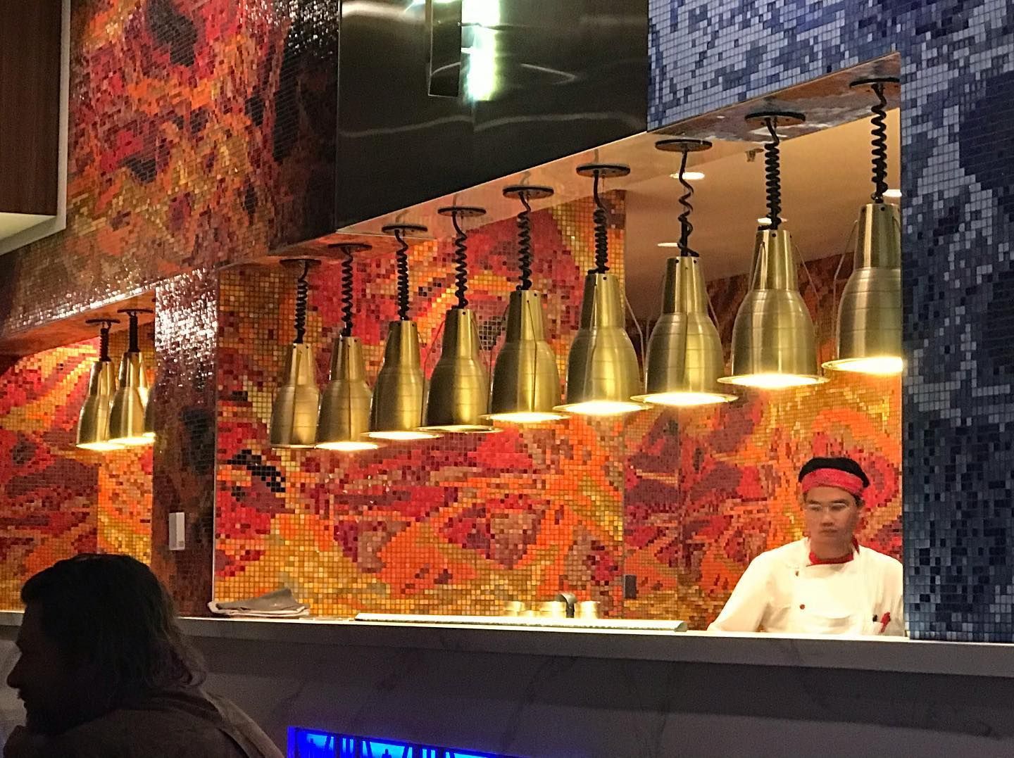 A chef works at a counter in front of a vibrant red mosaic wall, illuminated by a row of industrial brass pendant lights.