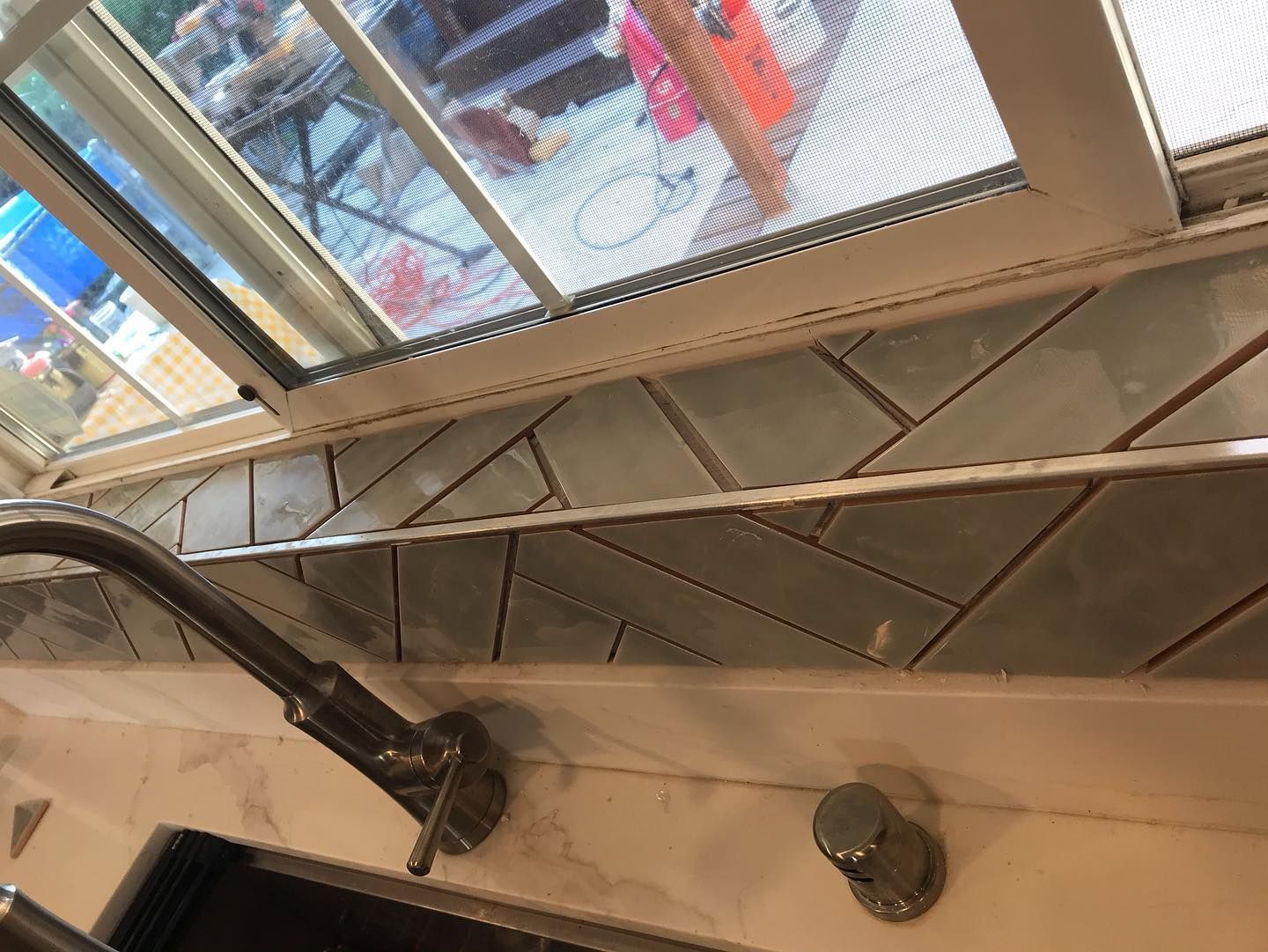 A kitchen window sill featuring gray, herringbone-patterned subway tiles next to a metal faucet and sink fixture.