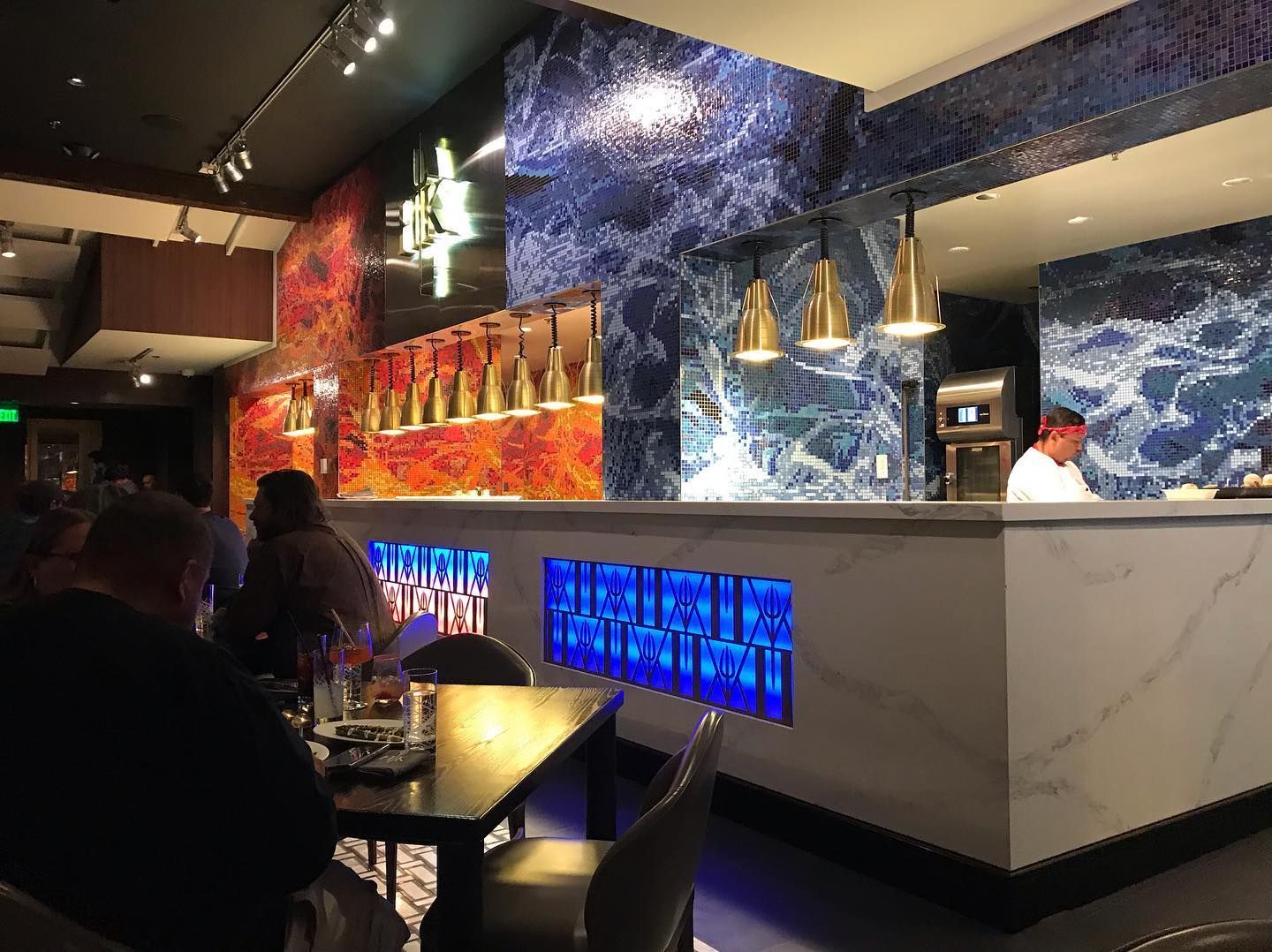 Modern restaurant interior featuring a bar with blue marble tiling, ambient pendant lighting, and diners at tables.