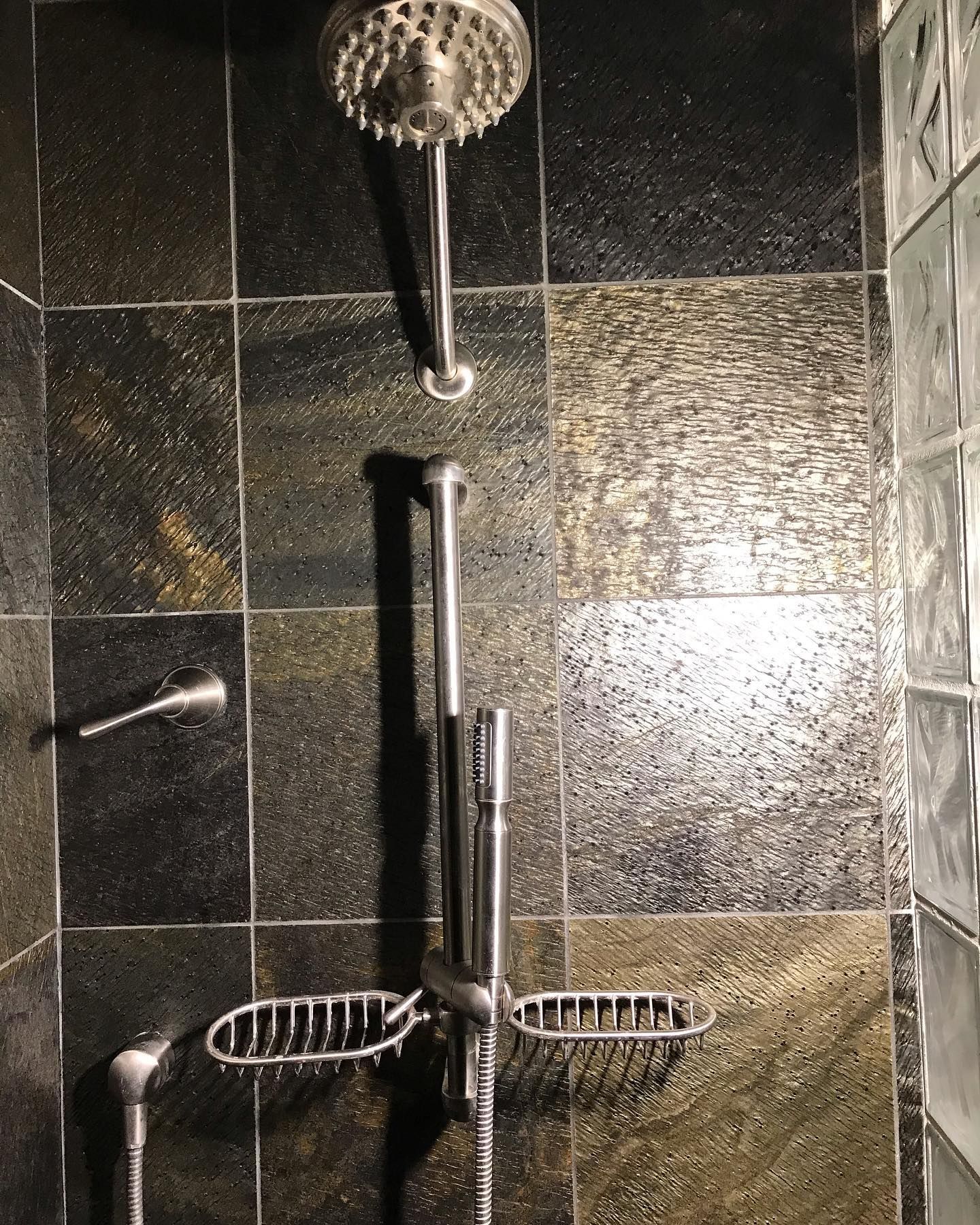 A shower stall featuring dark, textured stone tile walls, a wall-mounted showerhead, and a slide-bar handheld shower unit.