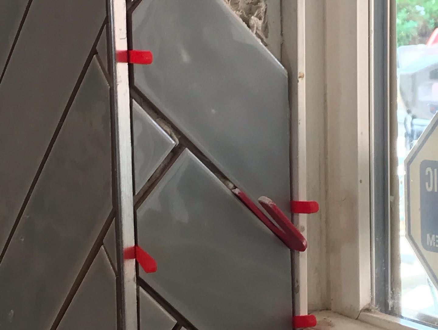 Gray rectangular tiles are being installed in a herringbone pattern on a wall next to a window using red leveling spacers.