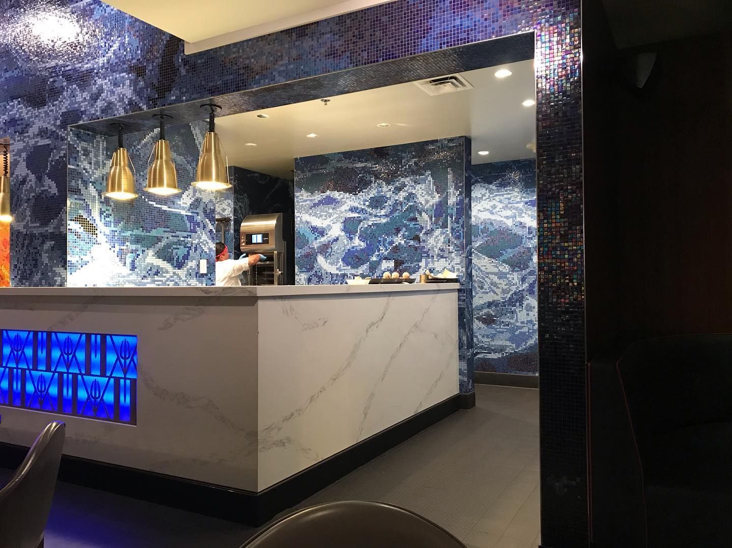 A modern bar counter with white marble, blue mosaic tile walls, and pendant lighting in a dimly lit, contemporary space.