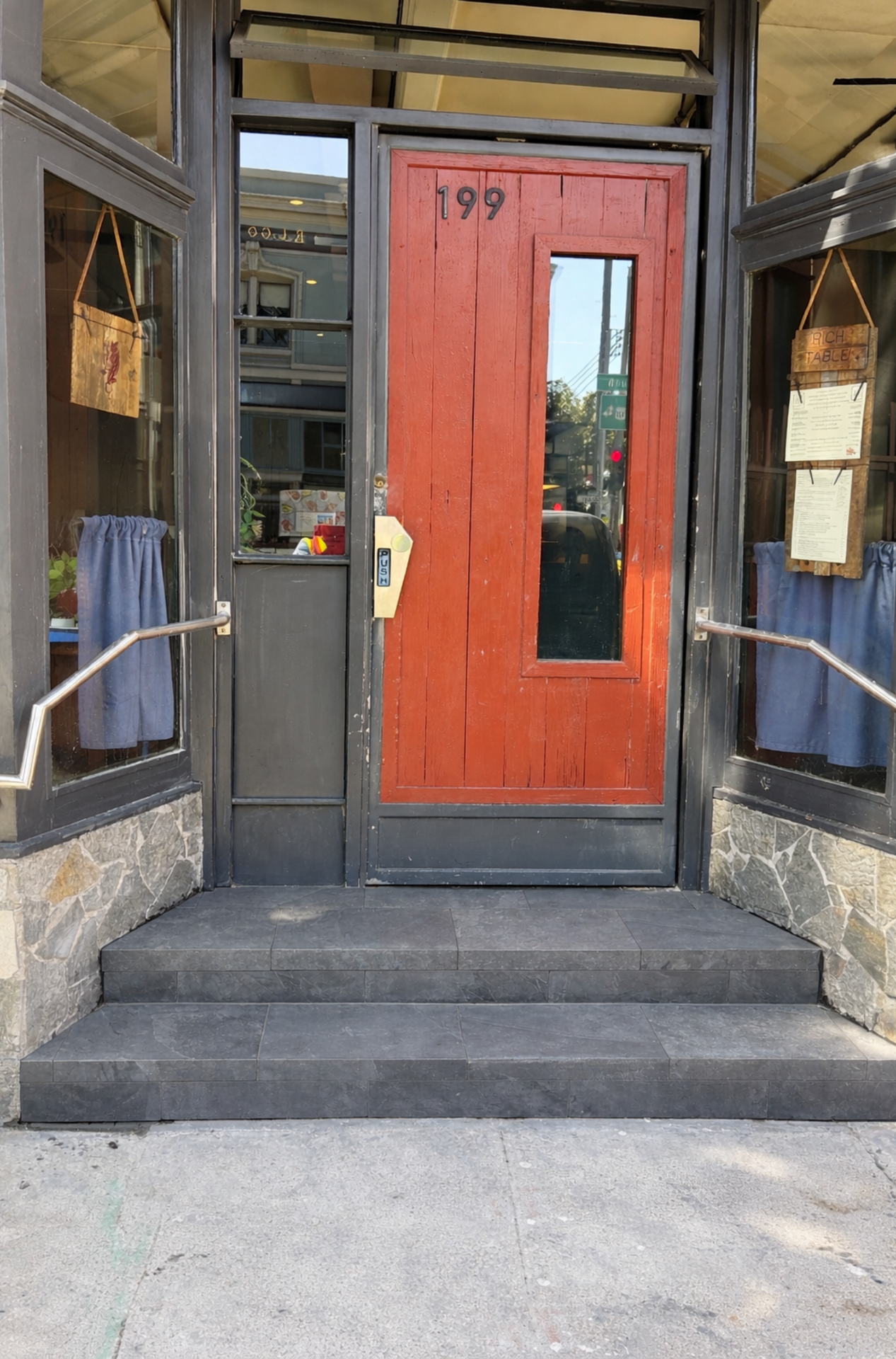 Red storefront door with glass panel and the number 199, framed by stone steps and blue curtains.