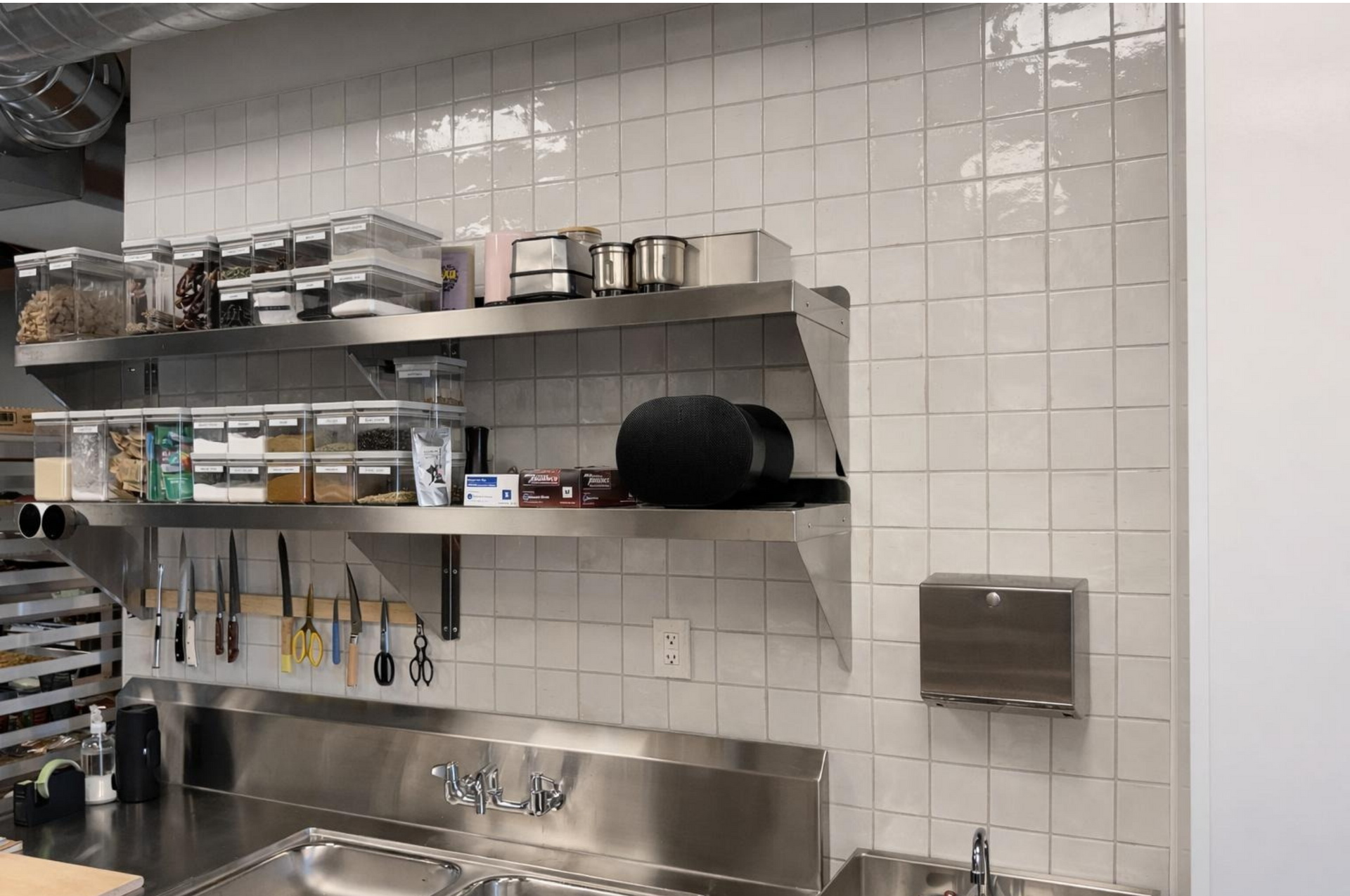 Stainless steel kitchen sink and shelves with pots, jars, utensils, and dish storage against white tiled wall