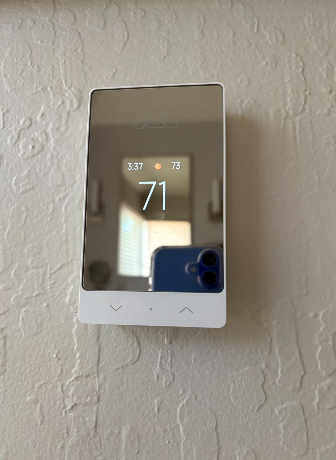 Wall-mounted digital thermostat on textured wall, displaying 71° and reflecting a blue phone at the bottom.
