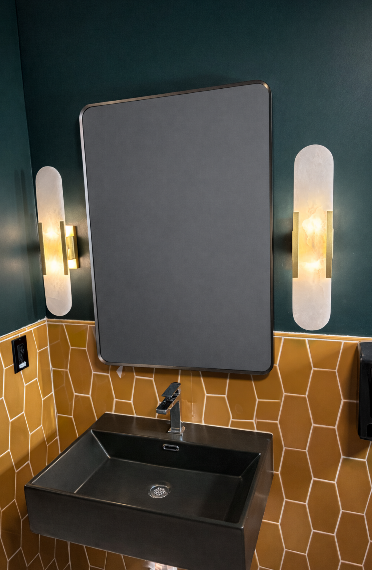 Modern bathroom vanity with black sink, rectangular mirror, warm wall sconces, and tan patterned tile backsplash