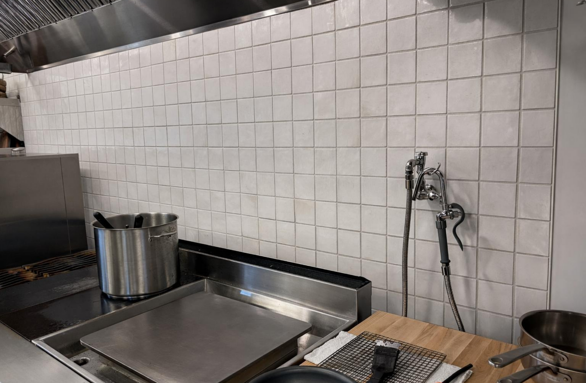 Commercial kitchen prep area with white tile wall, stainless steel counter, sink, and utensils