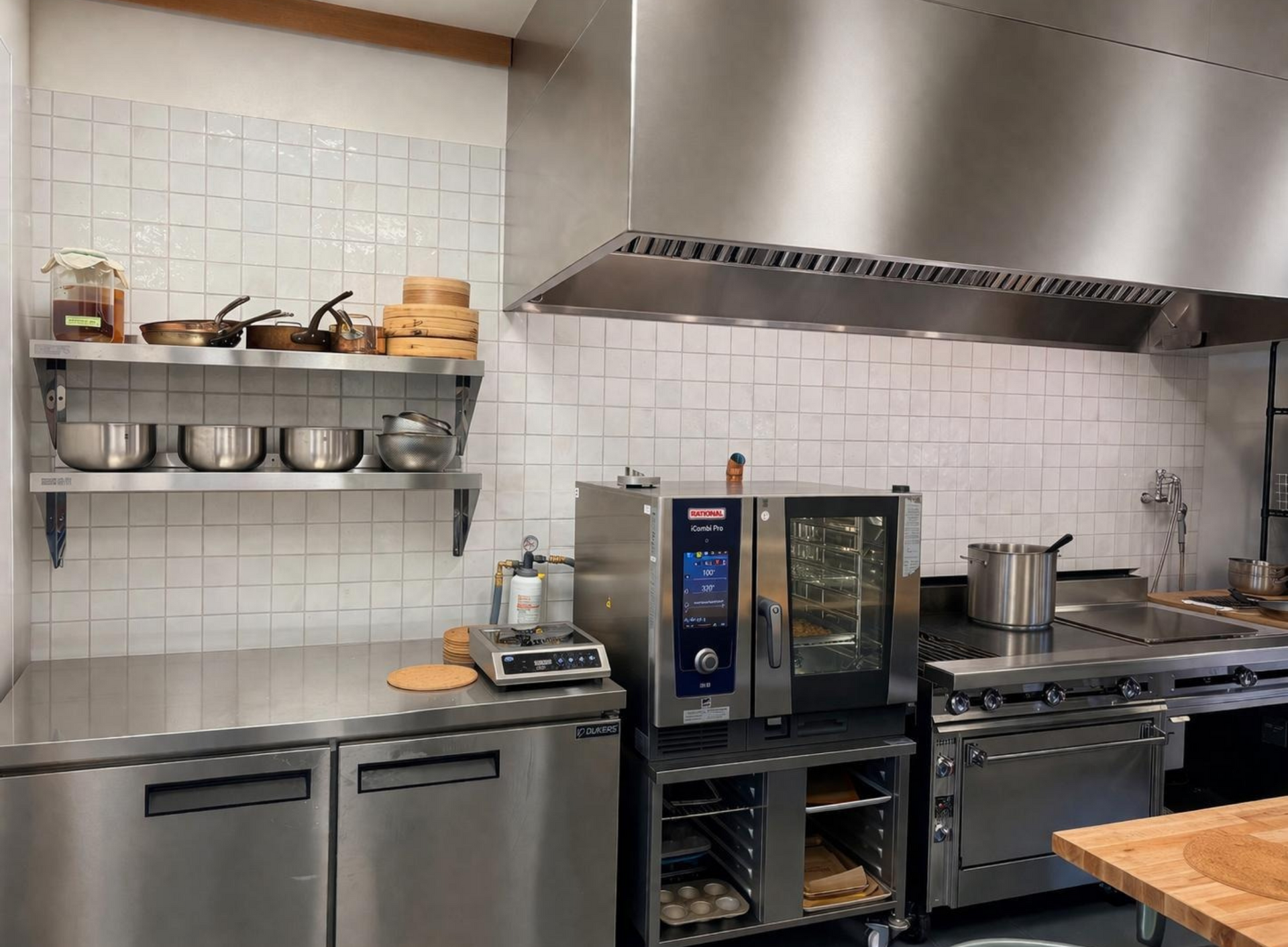 Commercial kitchen with stainless steel counters, shelves, oven, stovetop, and large vent hood.