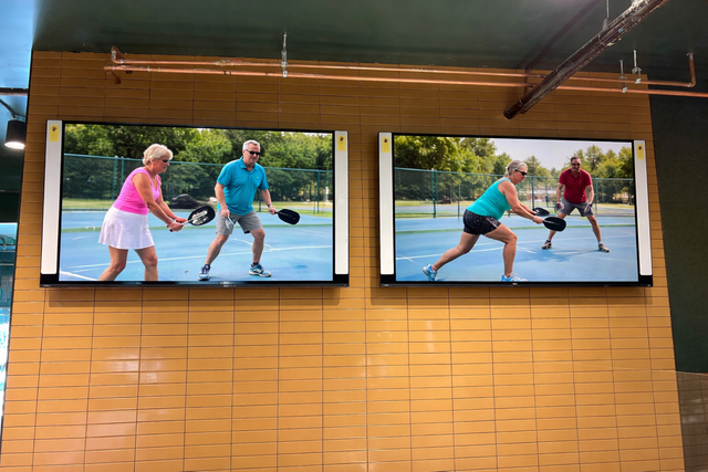 Two TV screens on a wooden wall show people playing pickleball indoors.