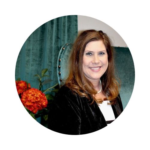 Smiling woman in a black jacket posed indoors beside teal curtains and orange flowers