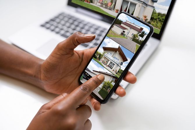 Hands holding a smartphone displaying images of houses, with a laptop visible in the background on a white surface.