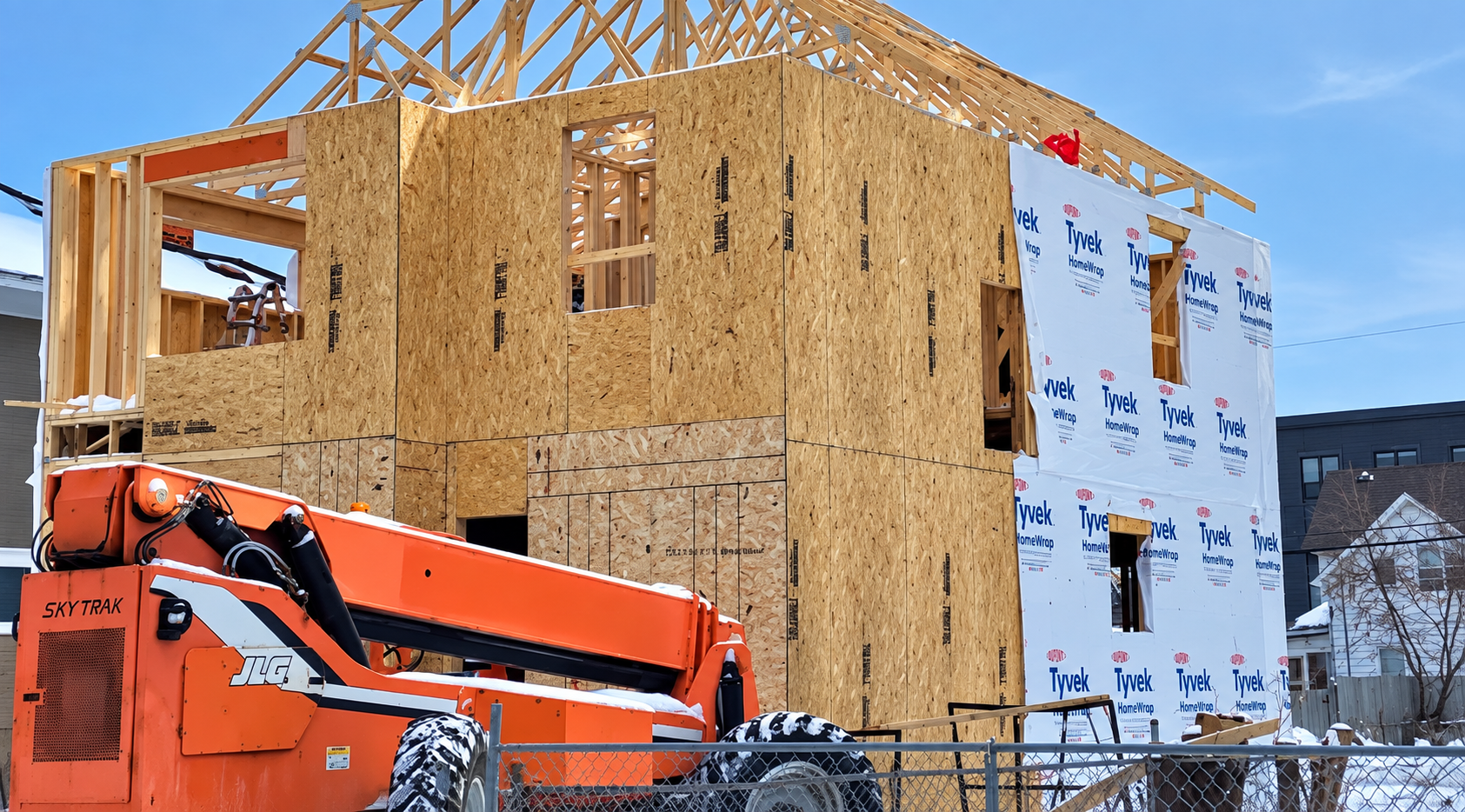 Orange construction lift beside a partially built house with exposed wood framing and wrapped exterior walls.
