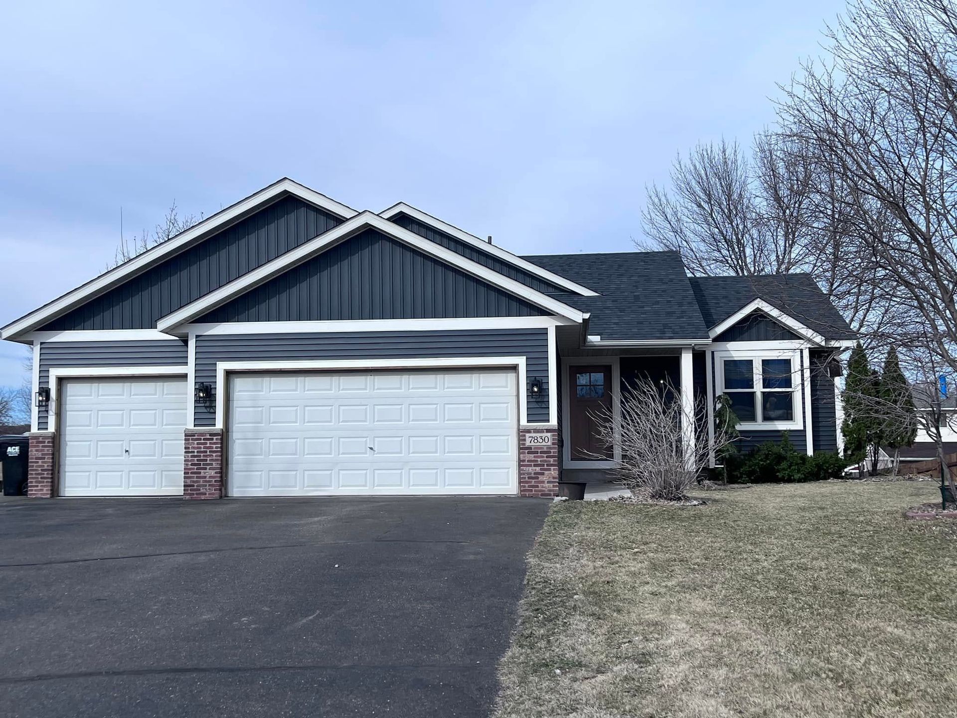 Suburban house with dark siding, three-car garage, and a wide driveway under a cloudy sky