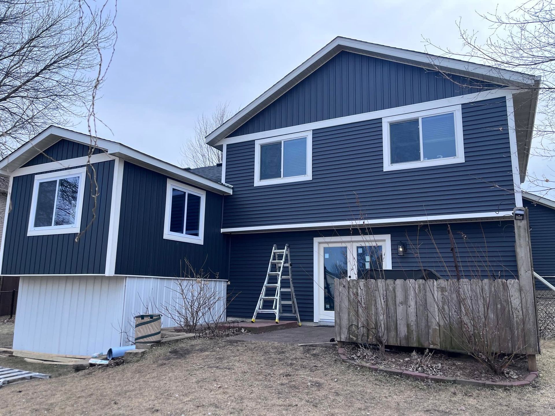 Blue two-story house with white lower siding, small attached shed, and a ladder on a dirt lot