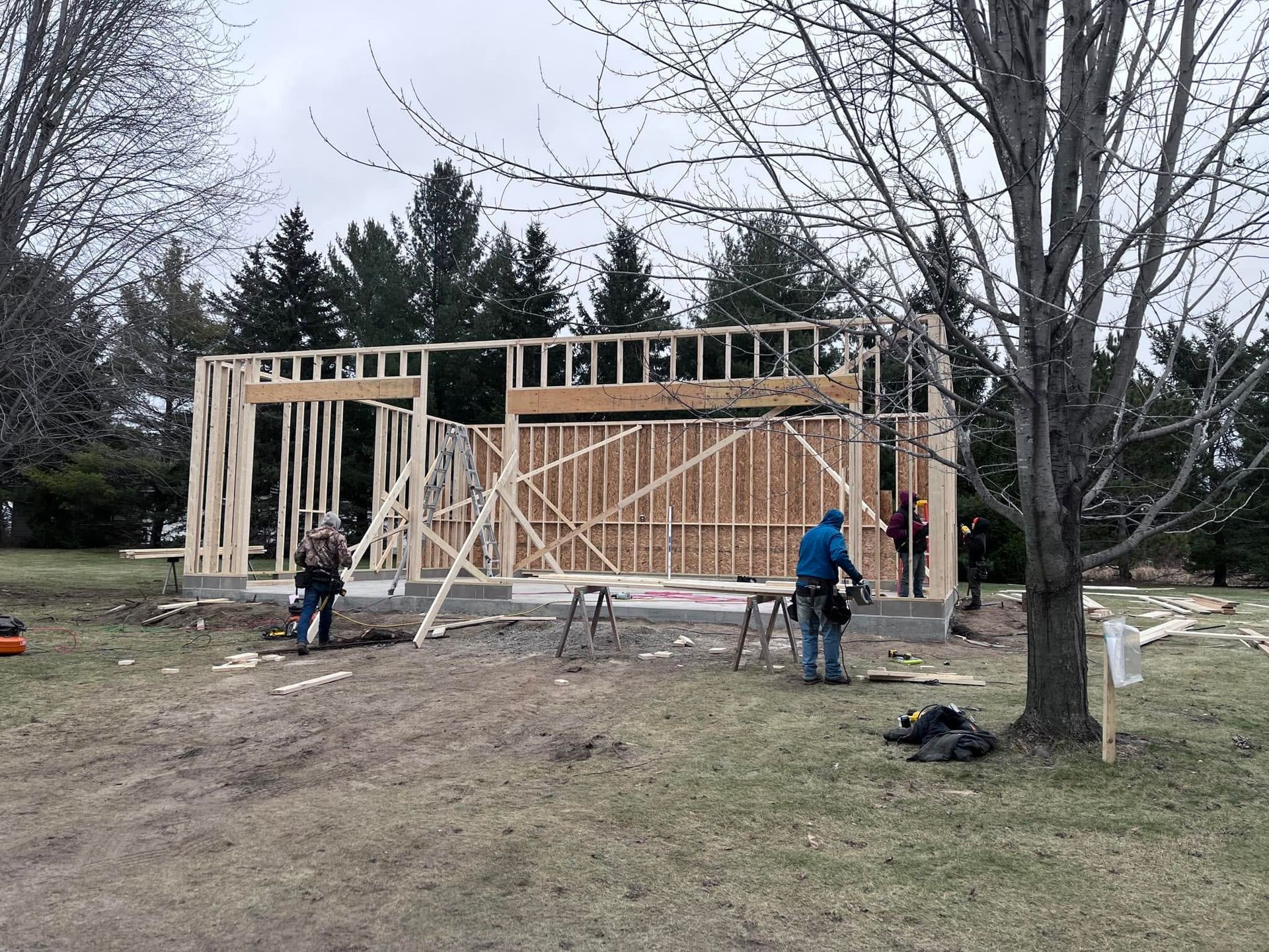 Workers framing a wooden house under construction in a bare yard, with trees in the background.