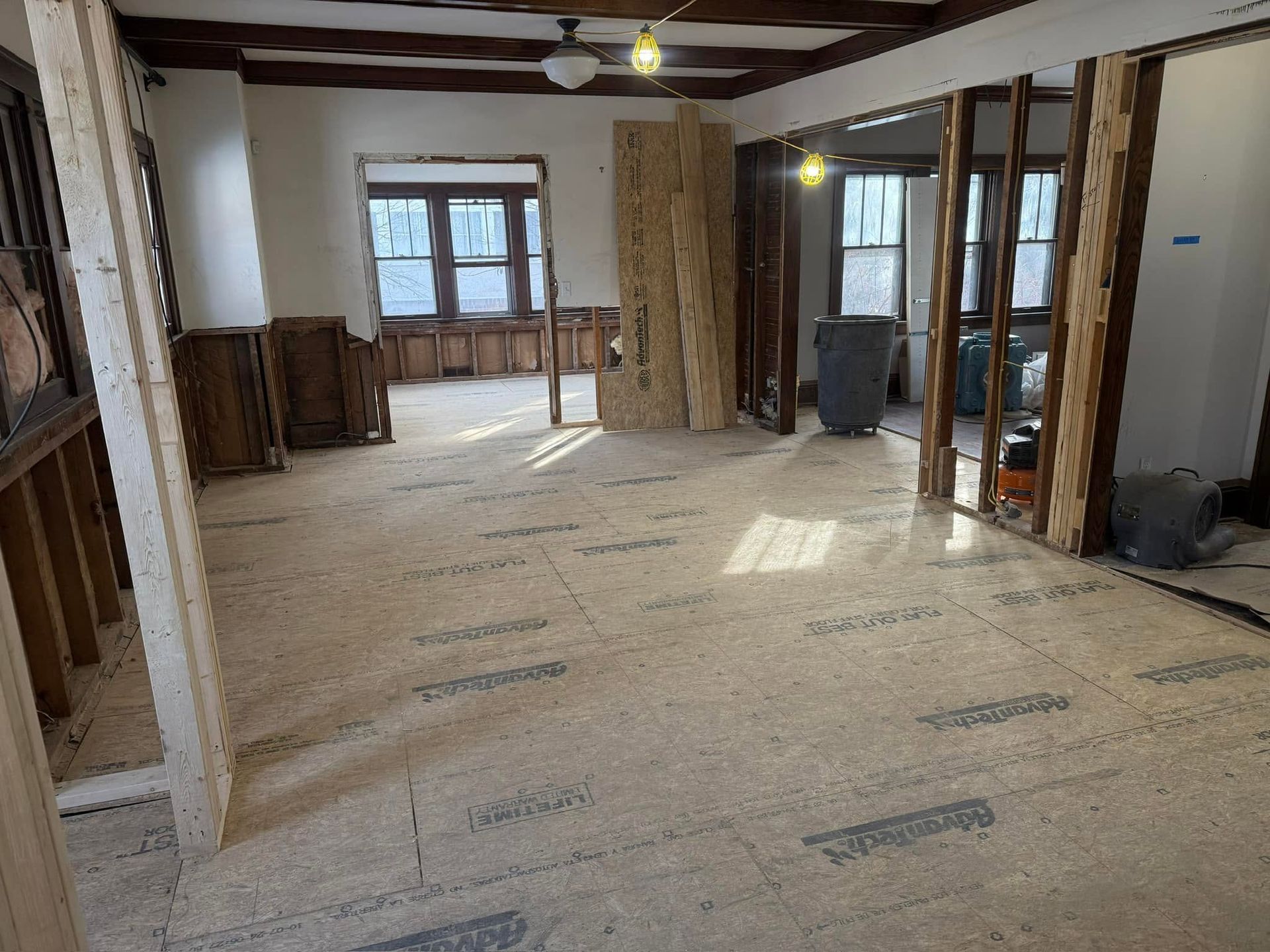 Unfinished room with exposed framing, plywood floor, and sunlight through windows and glass doors