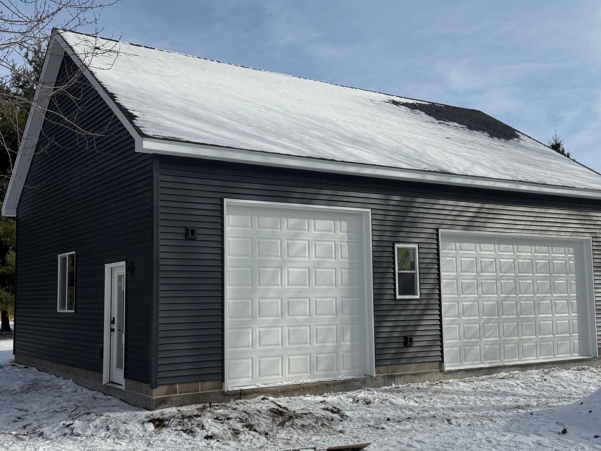 Black garage with two white doors and a snow-covered roof in winter.
