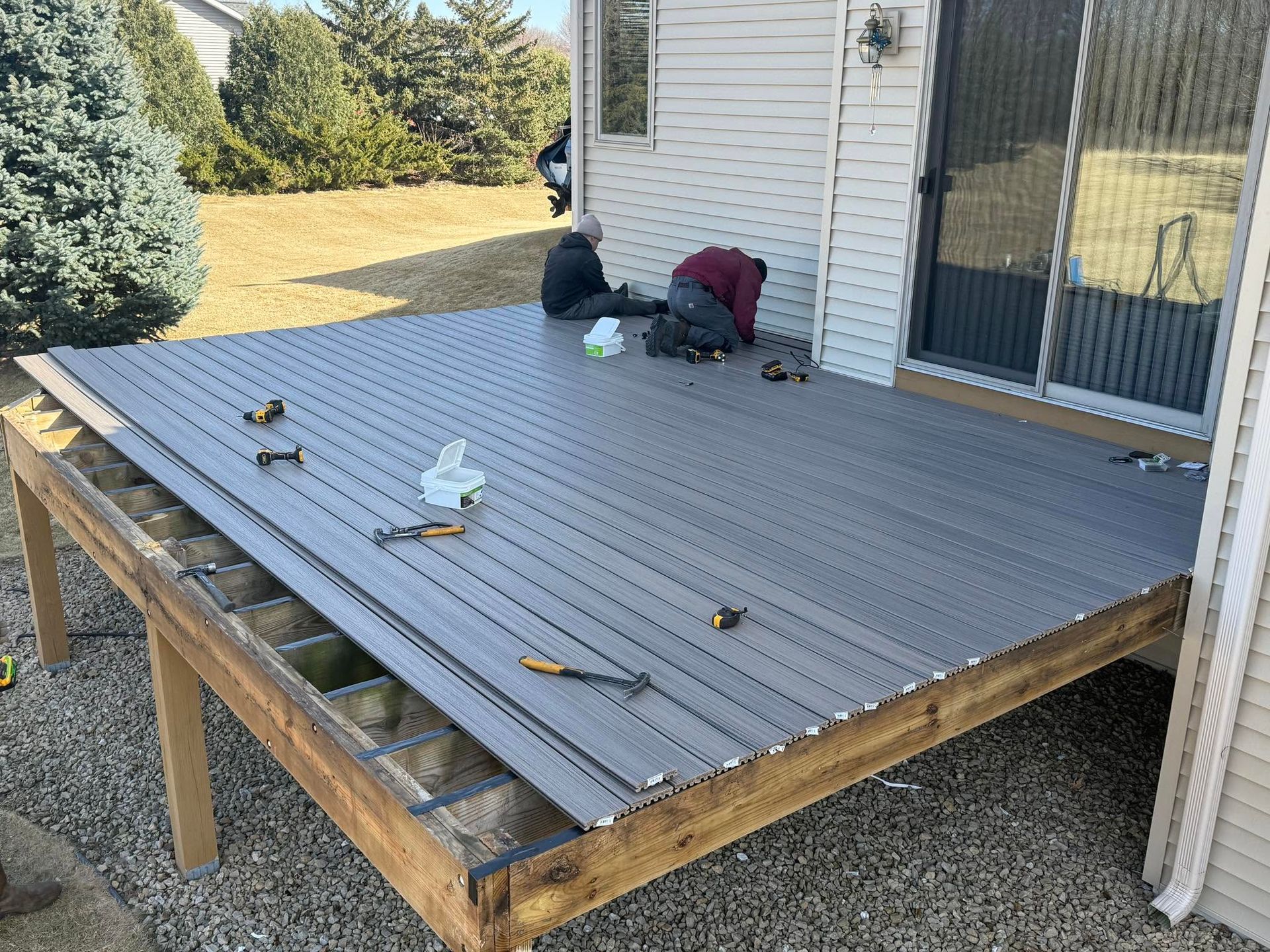 People working on a gray wooden deck beside a white house, with tools scattered around.