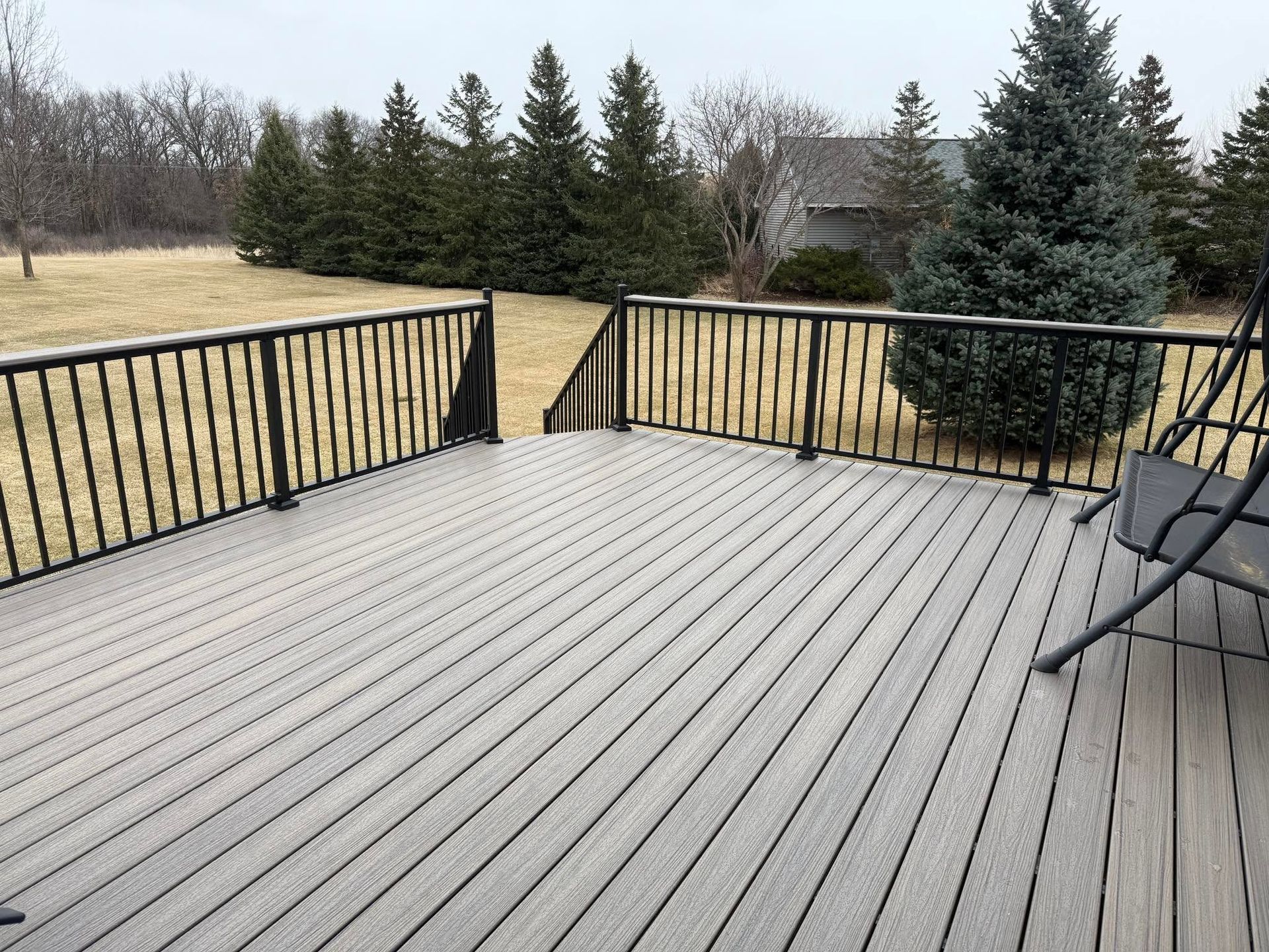 Wooden deck with black railing overlooking a snowy yard and evergreen trees