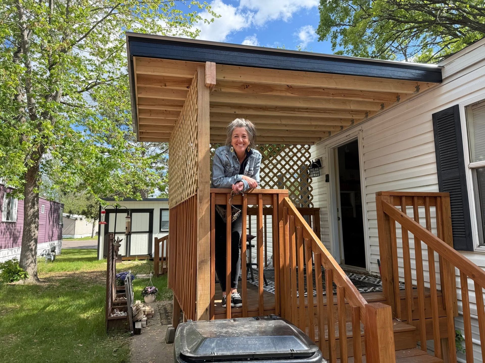 Person standing on a wooden porch with stairs under a covered roof, in a grassy yard.