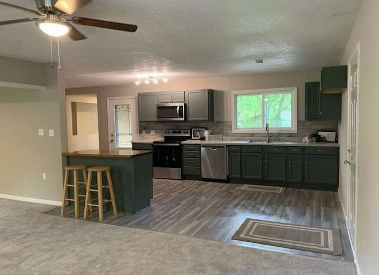 Open-concept kitchen and living area with dark cabinets, stainless appliances, island stools, and a ceiling fan.