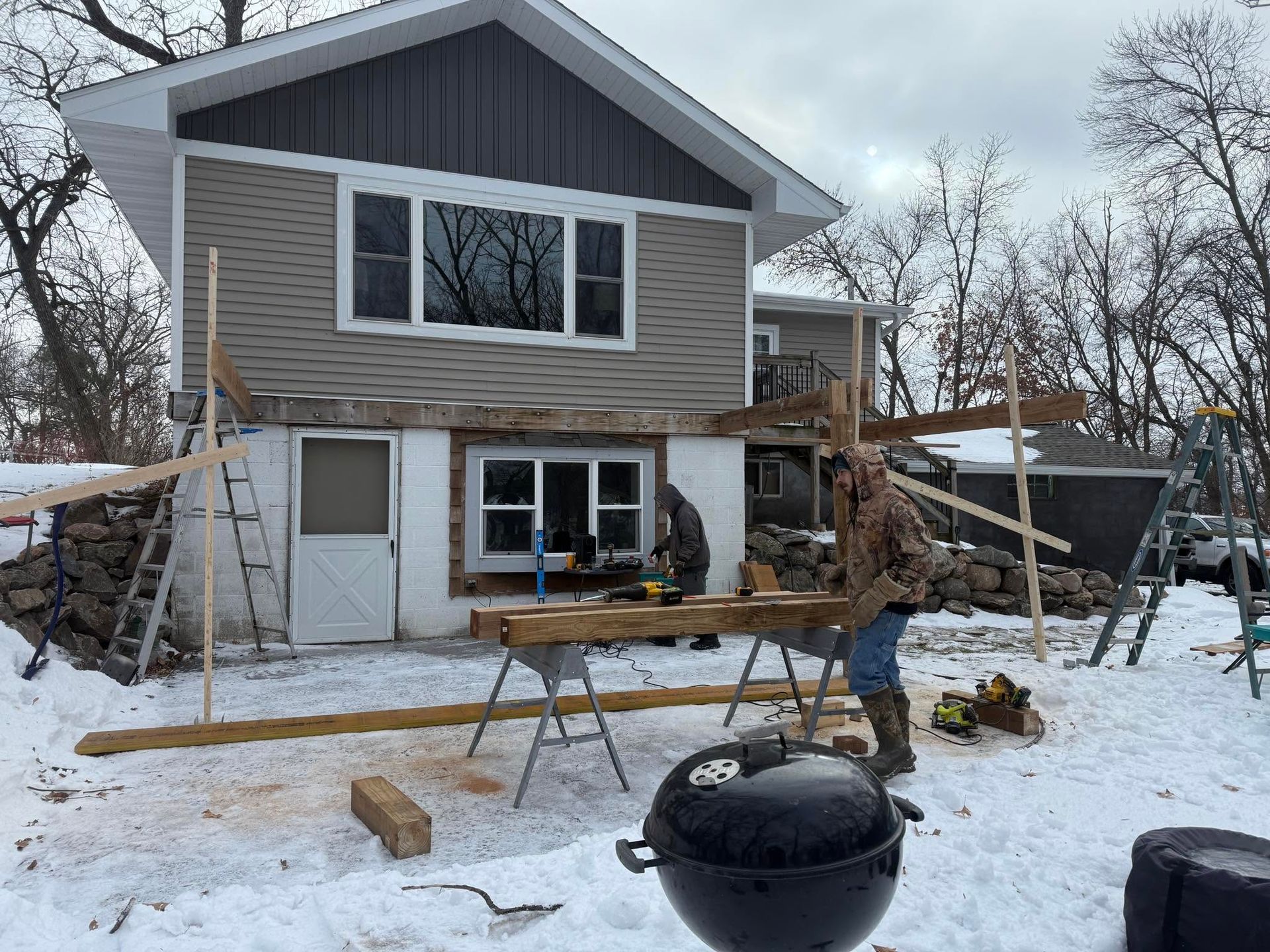 Snowy house under construction with a worker, ladders, and lumber in the yard