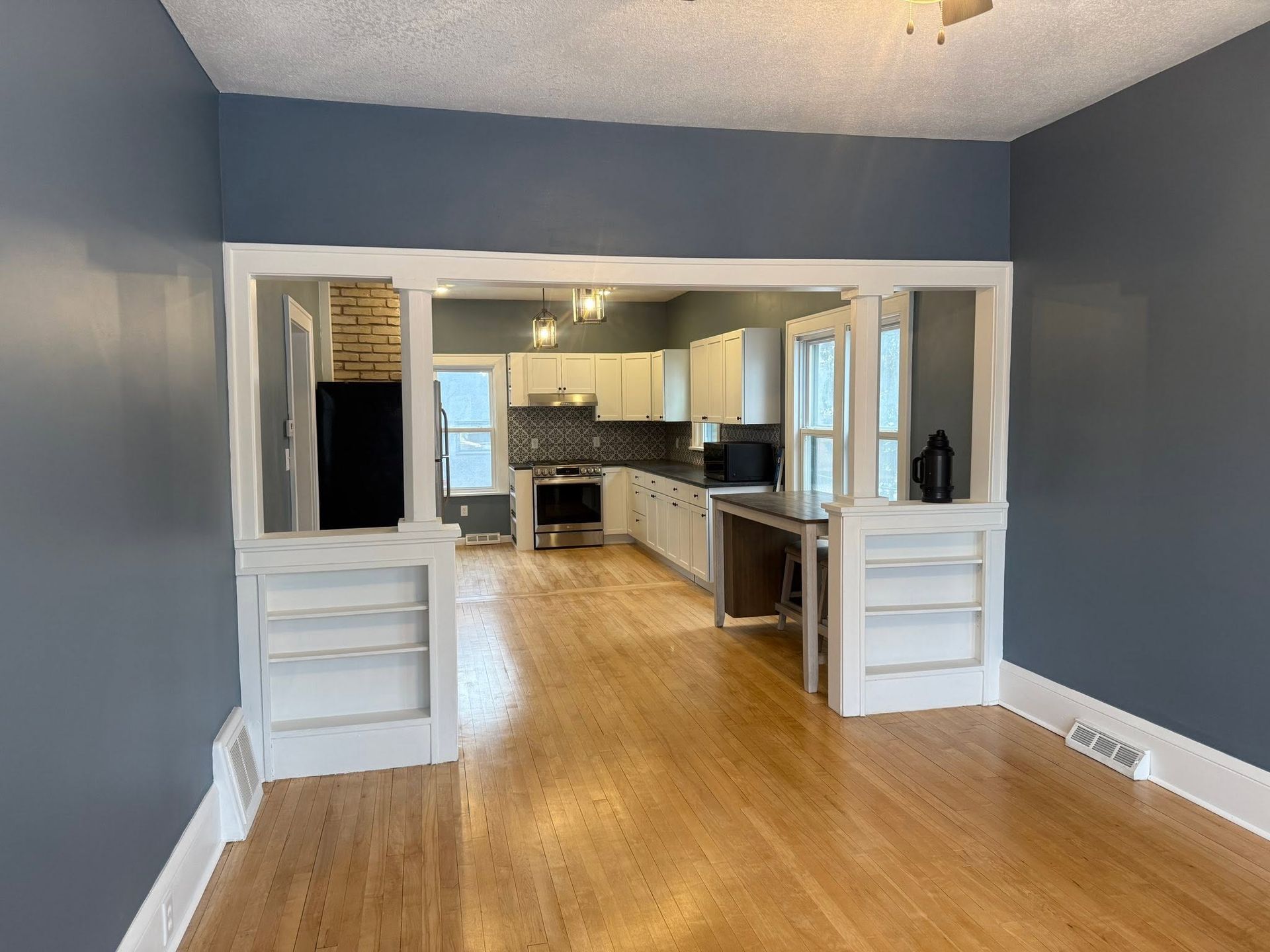 Empty room with blue walls, wood floor, and an open view into a bright kitchen.