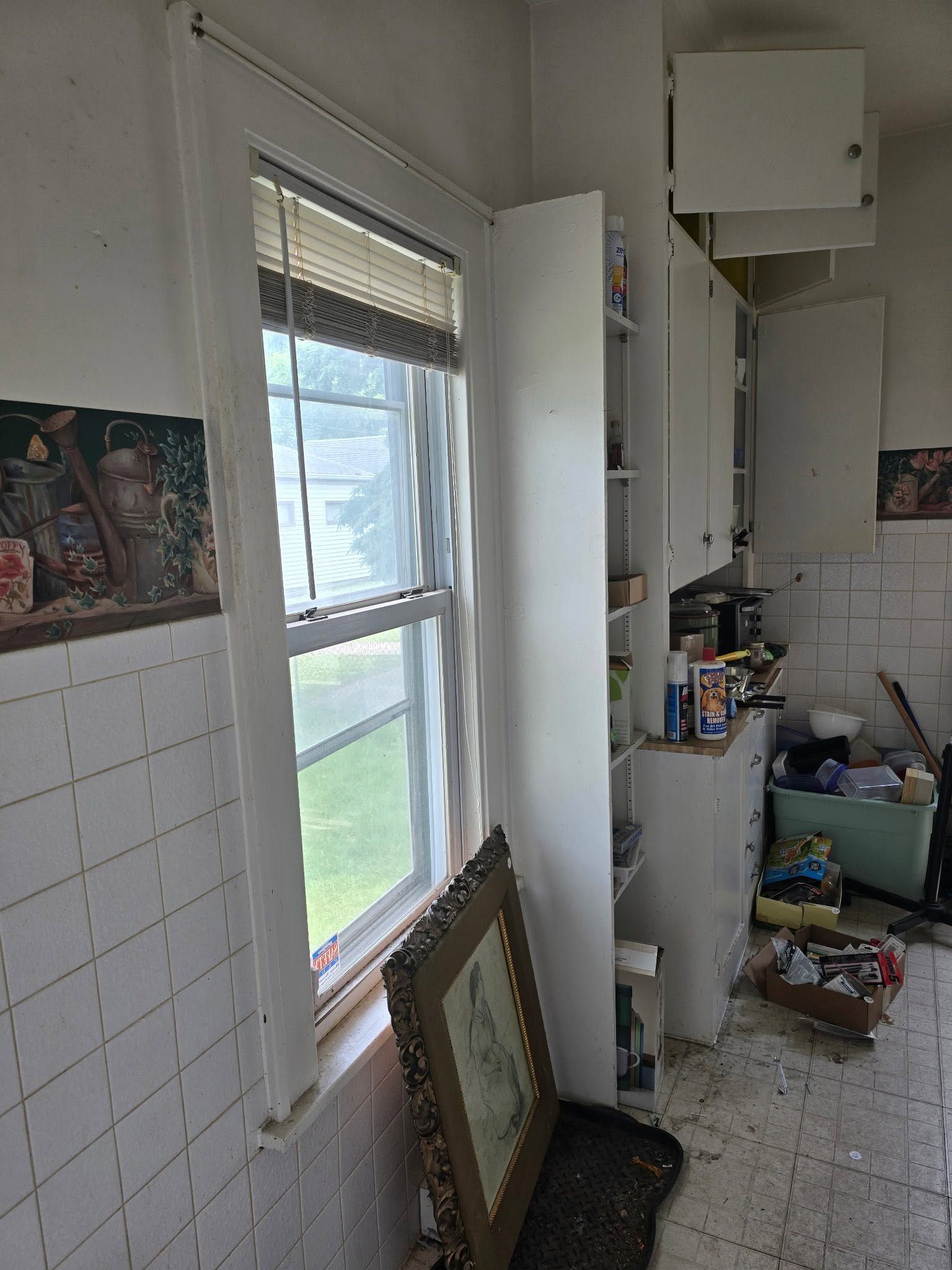 Narrow tiled kitchen with a window, open cabinet, and cleaning supplies on the floor.