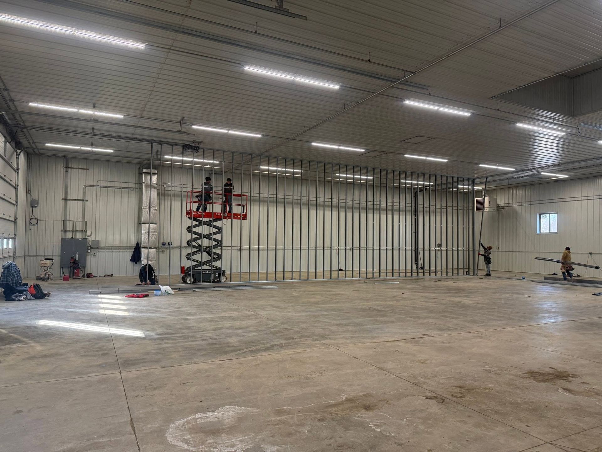 Empty industrial warehouse with concrete floor, metal wall framing, and a red scissor lift under bright ceiling lights