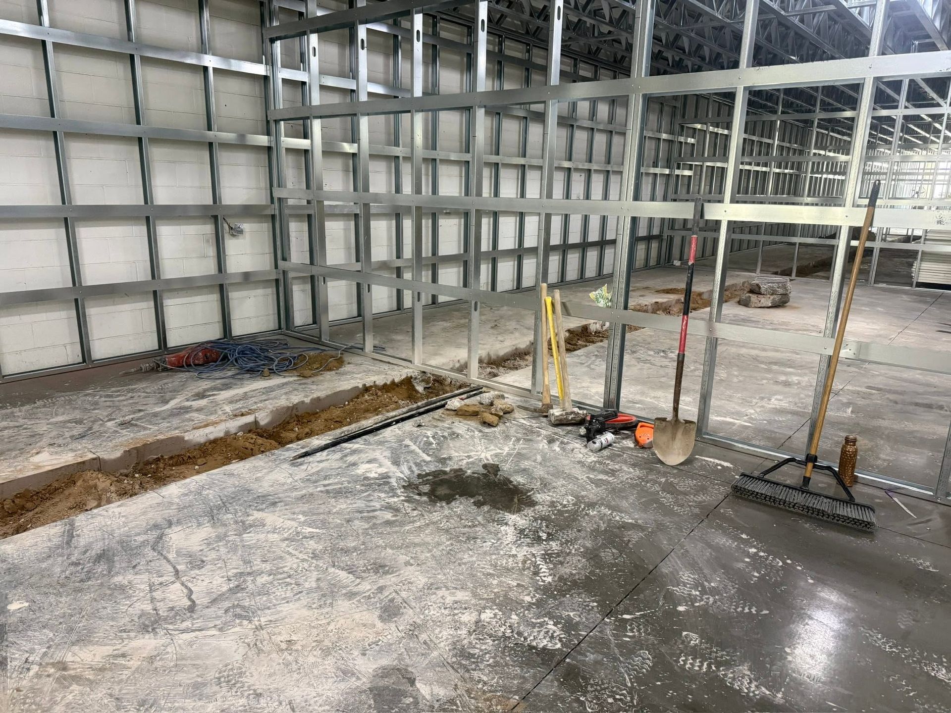 Construction site interior with wet concrete floor, metal wall framing, and tools near a trench.