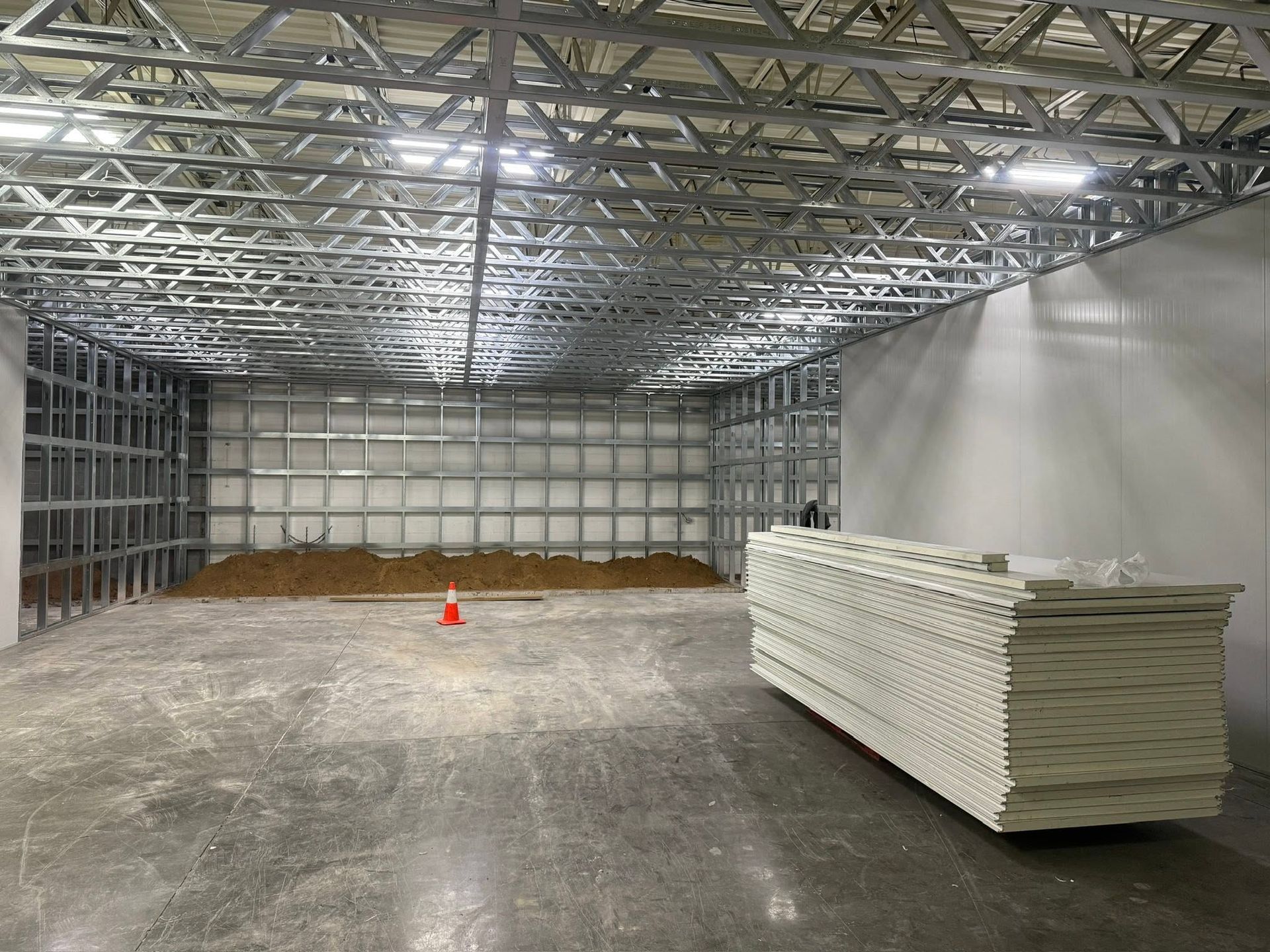 Empty warehouse with metal roof trusses, stacked white panels on the right, and an orange cone on the floor.