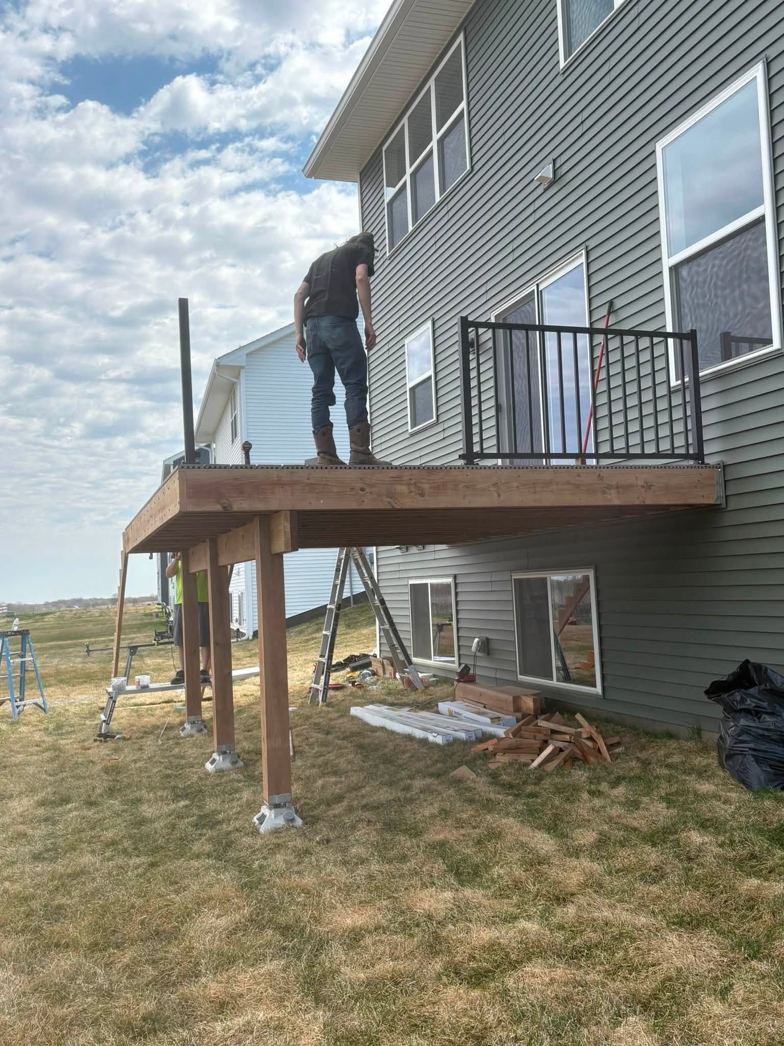 Person standing on a wooden deck attached to a gray house, with railing and grassy yard below.