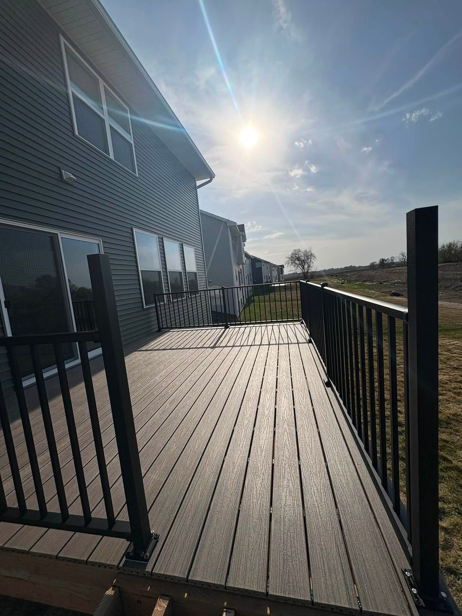 Sunlit wooden deck beside a house, with black metal railing and a bright sky over the water.