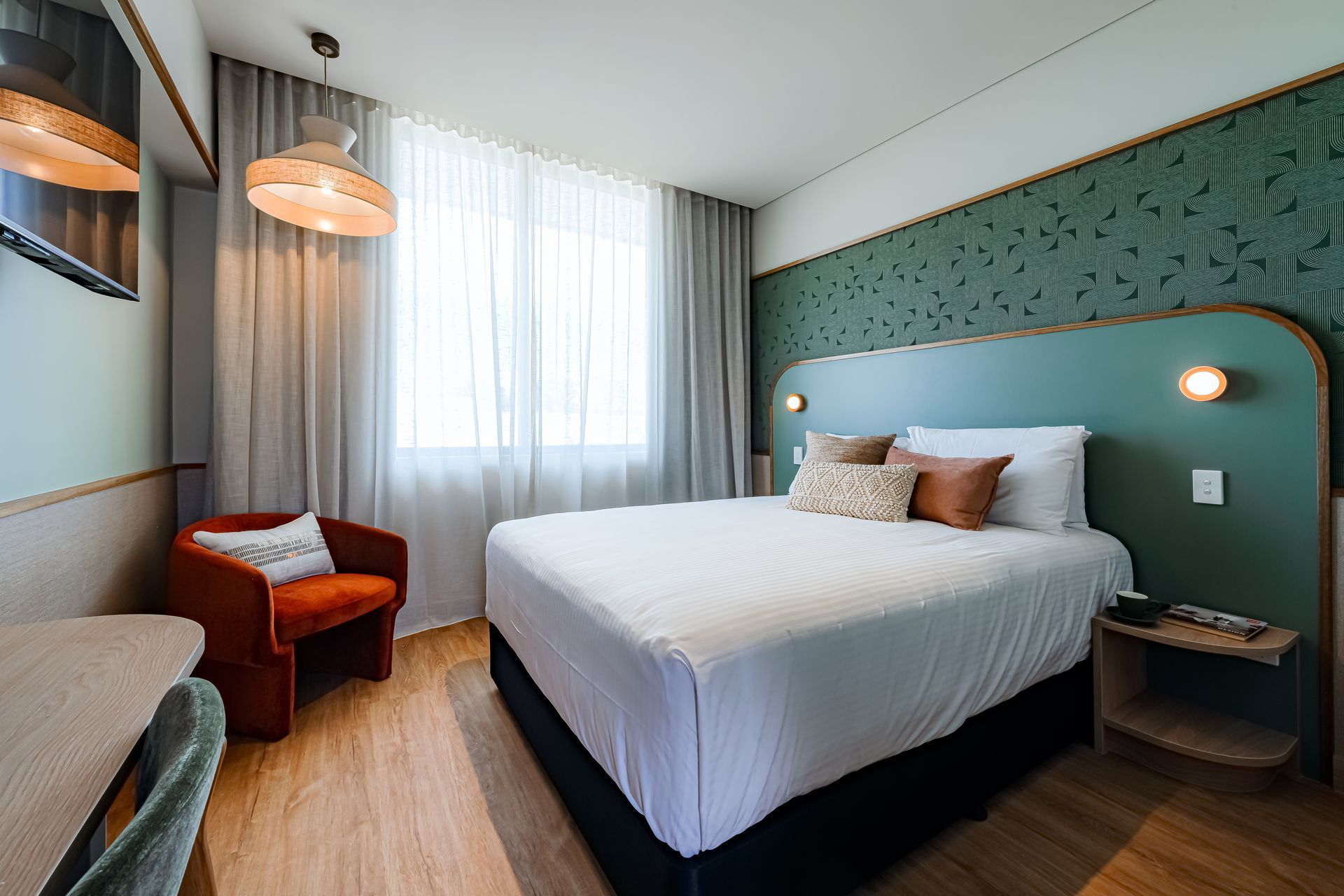 Modern hotel room with a bed, teal accent wall, window, chair, and warm pendant lights