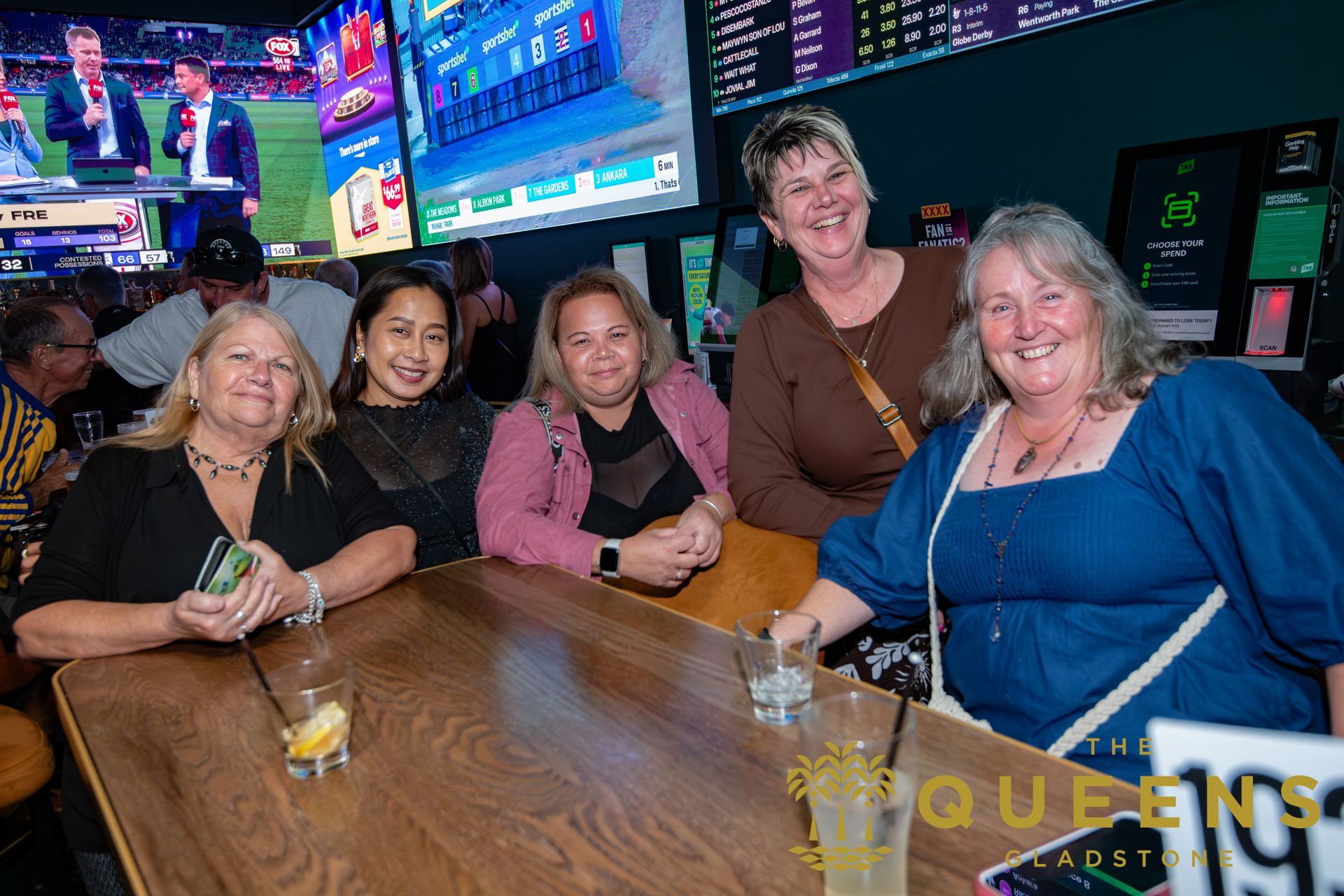 Five friends smiling at a bar table with drinks and TVs in the background