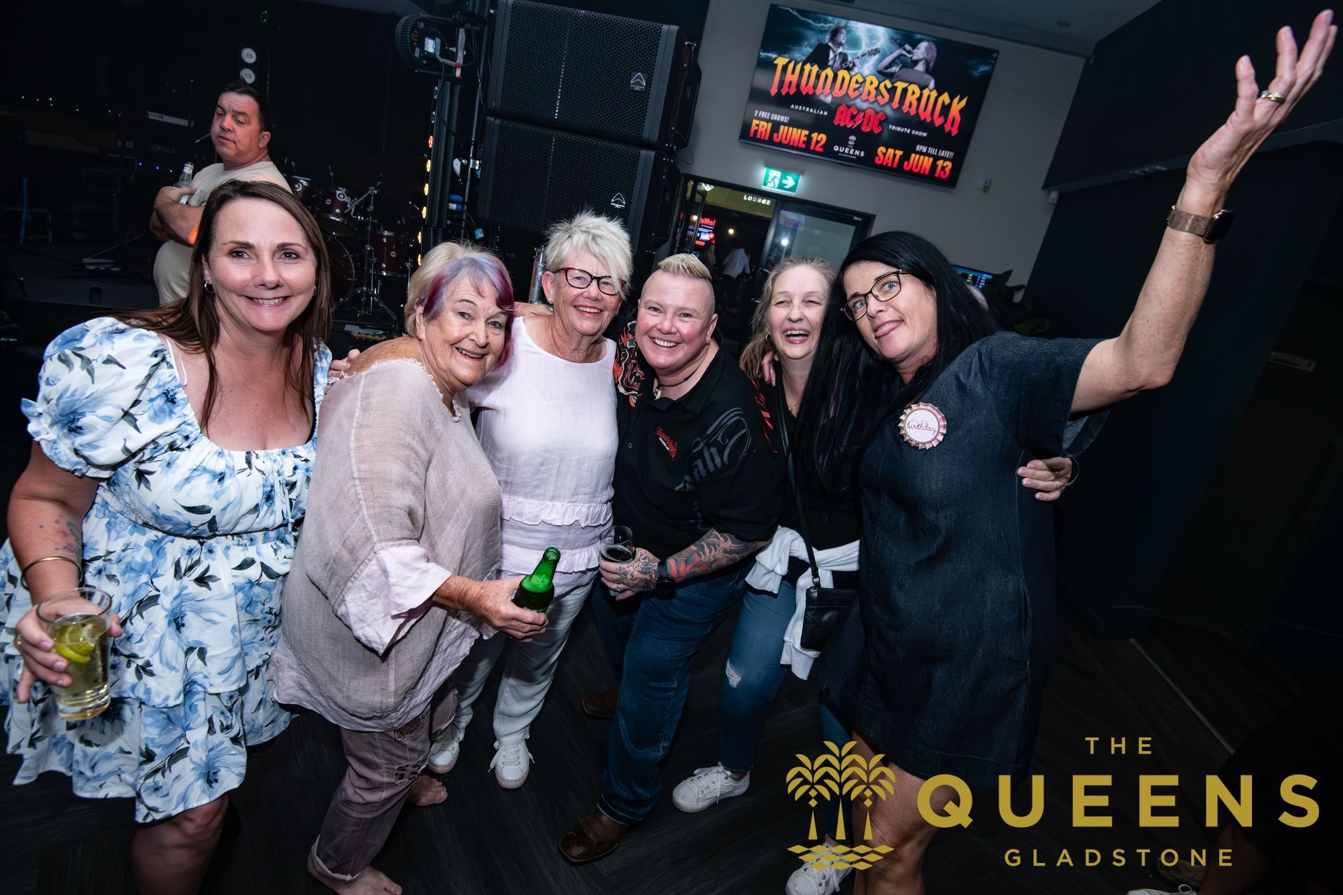 Six smiling friends posing indoors at a nightclub, one holding a green bottle, with 