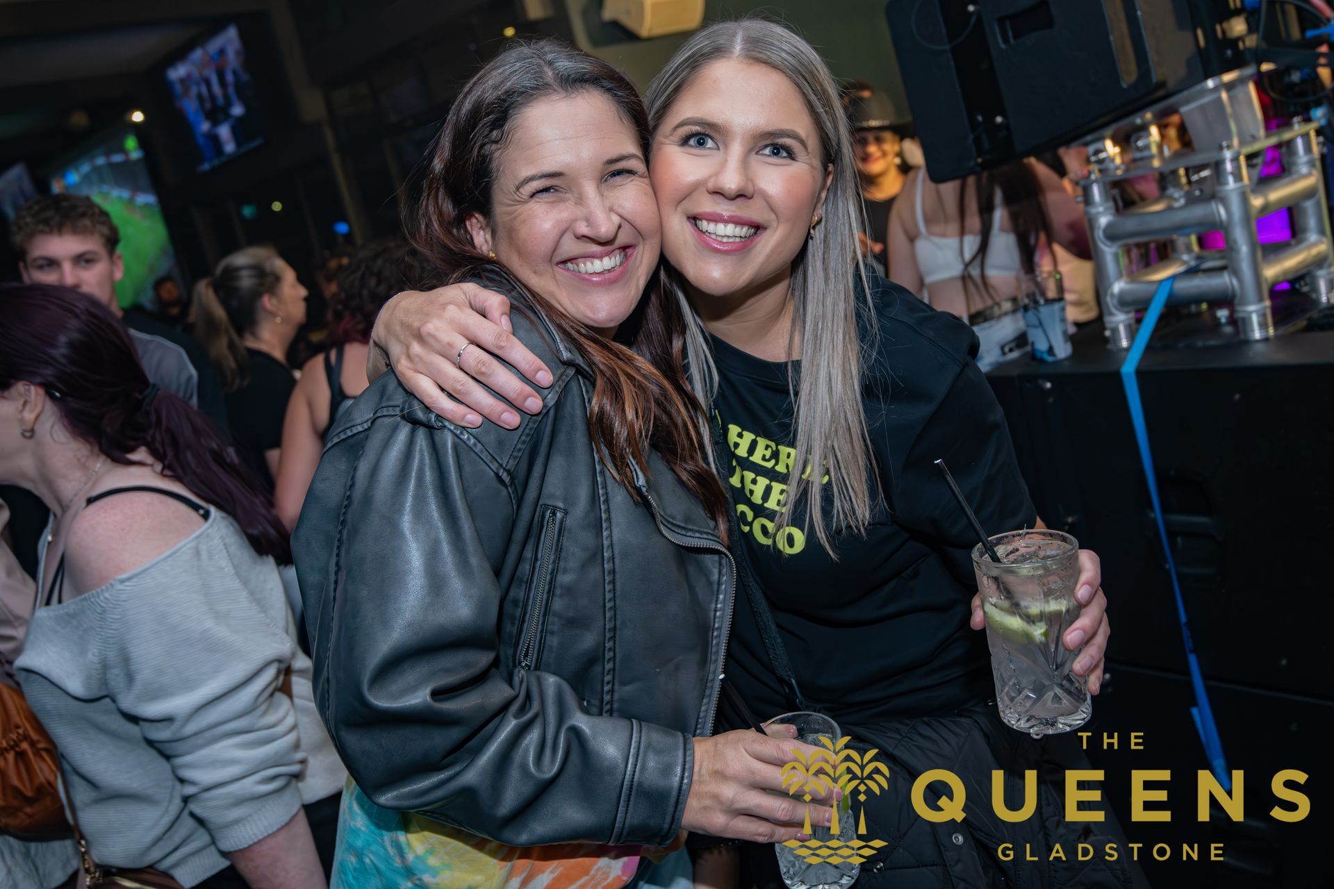 Two women smiling and hugging at a nightclub, one holding a drink, with neon lights and QUEENS Gladstone sign.